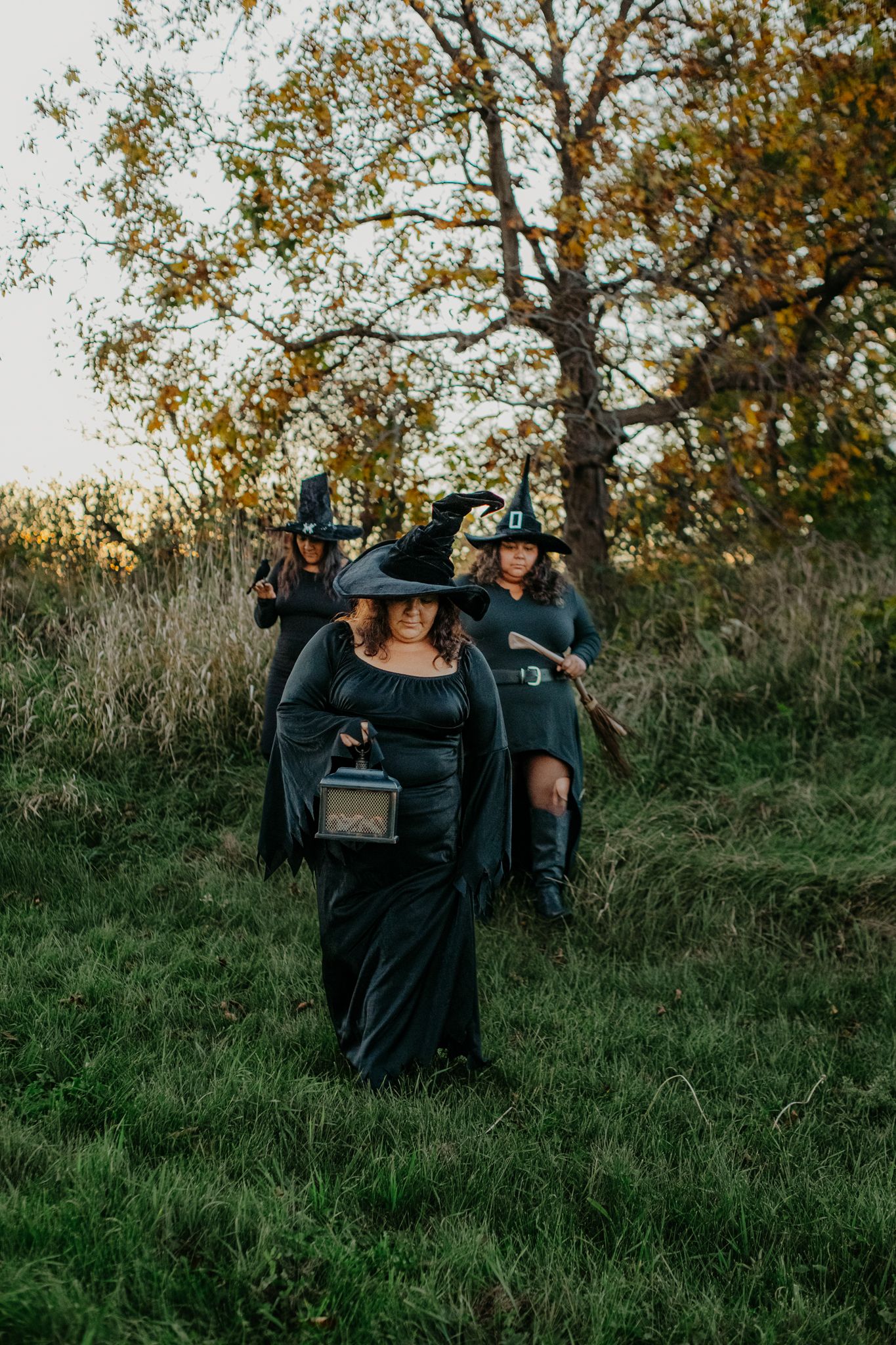 A group in dark clothing carrying lanterns walks through a grassy field at dusk with autumn trees in the background.