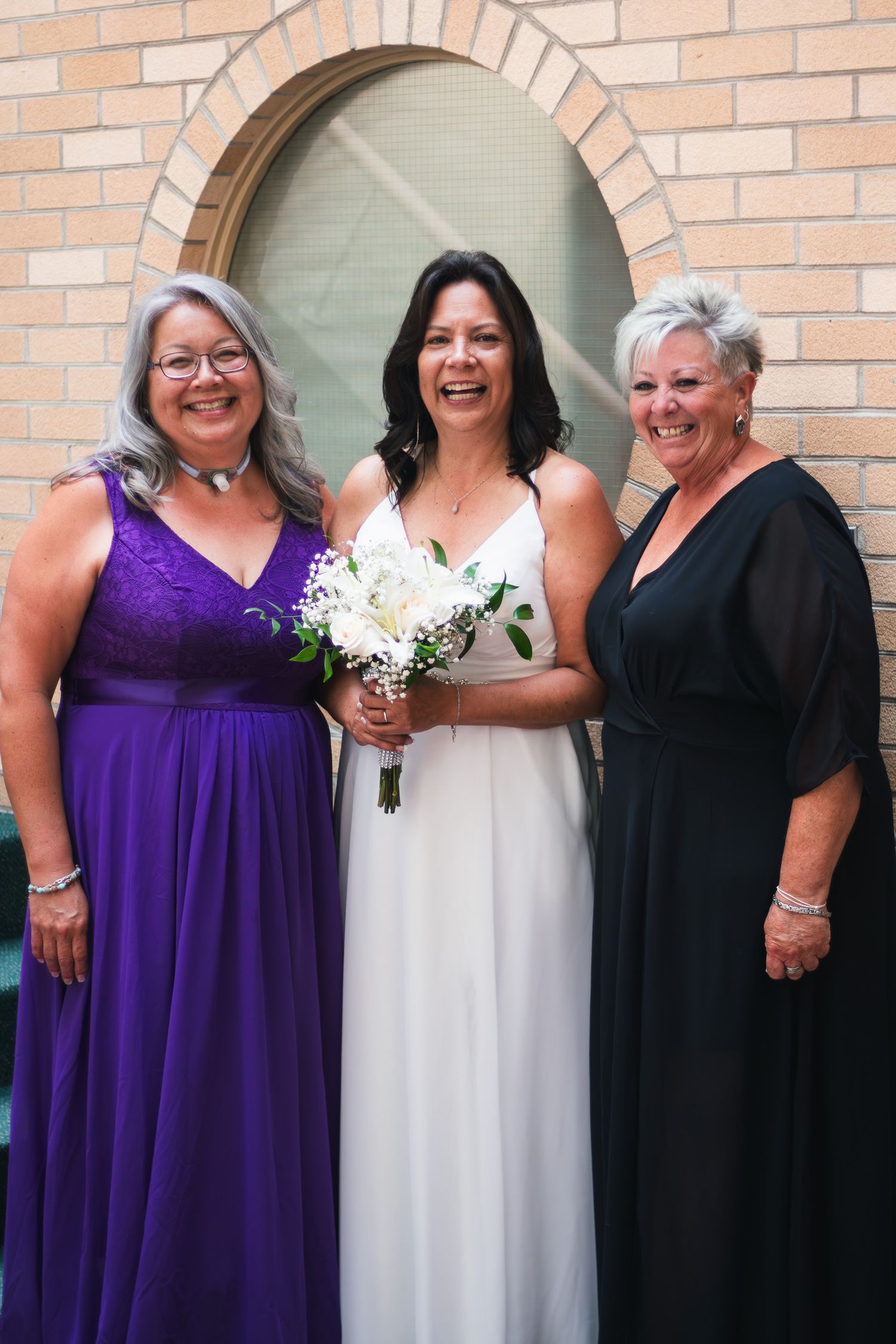 Bride in white gown laughing with two women in purple and black dresses holding white bouquet outside brick building.