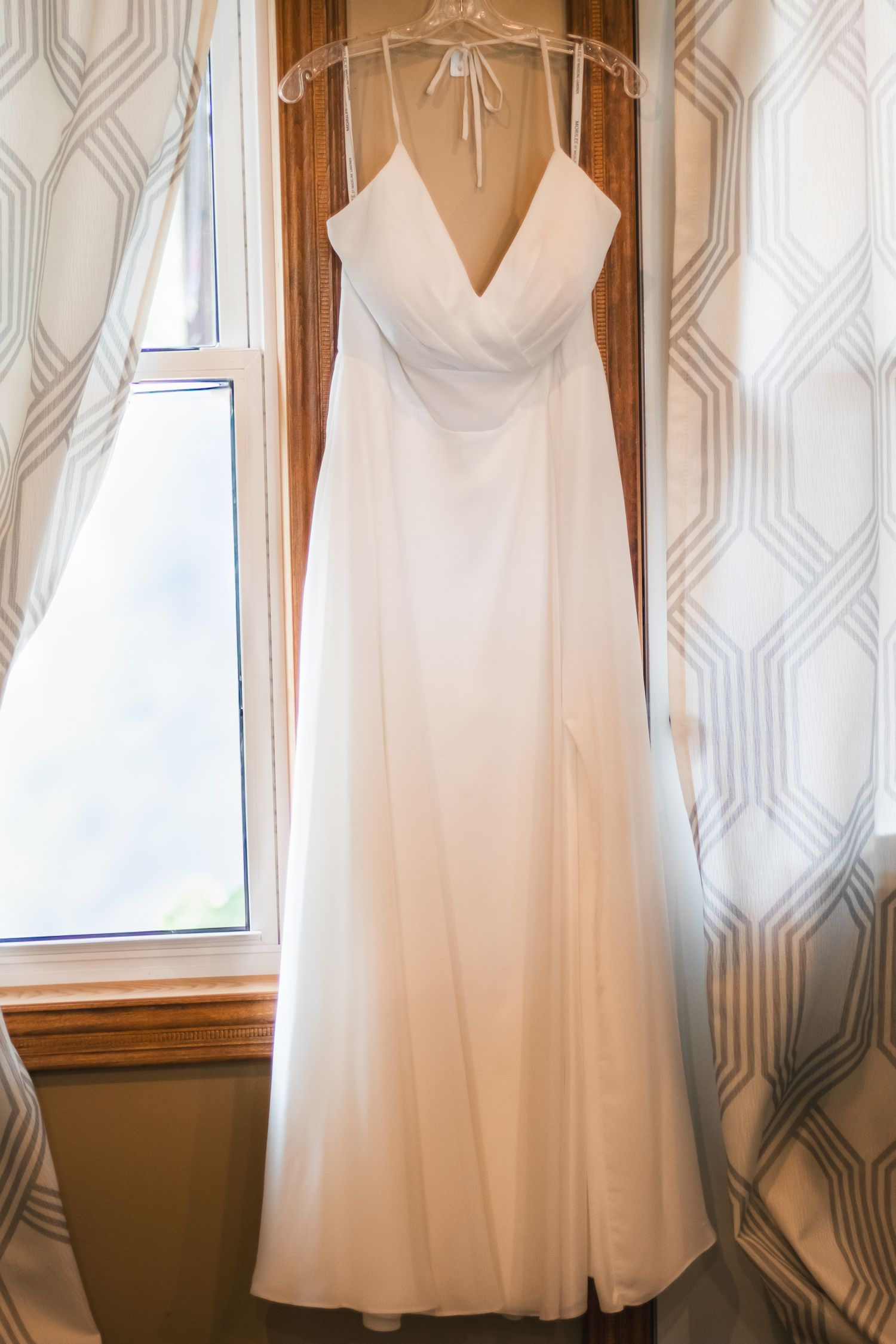 Elegant white V-neck spaghetti strap wedding gown hanging in window with striped curtains.