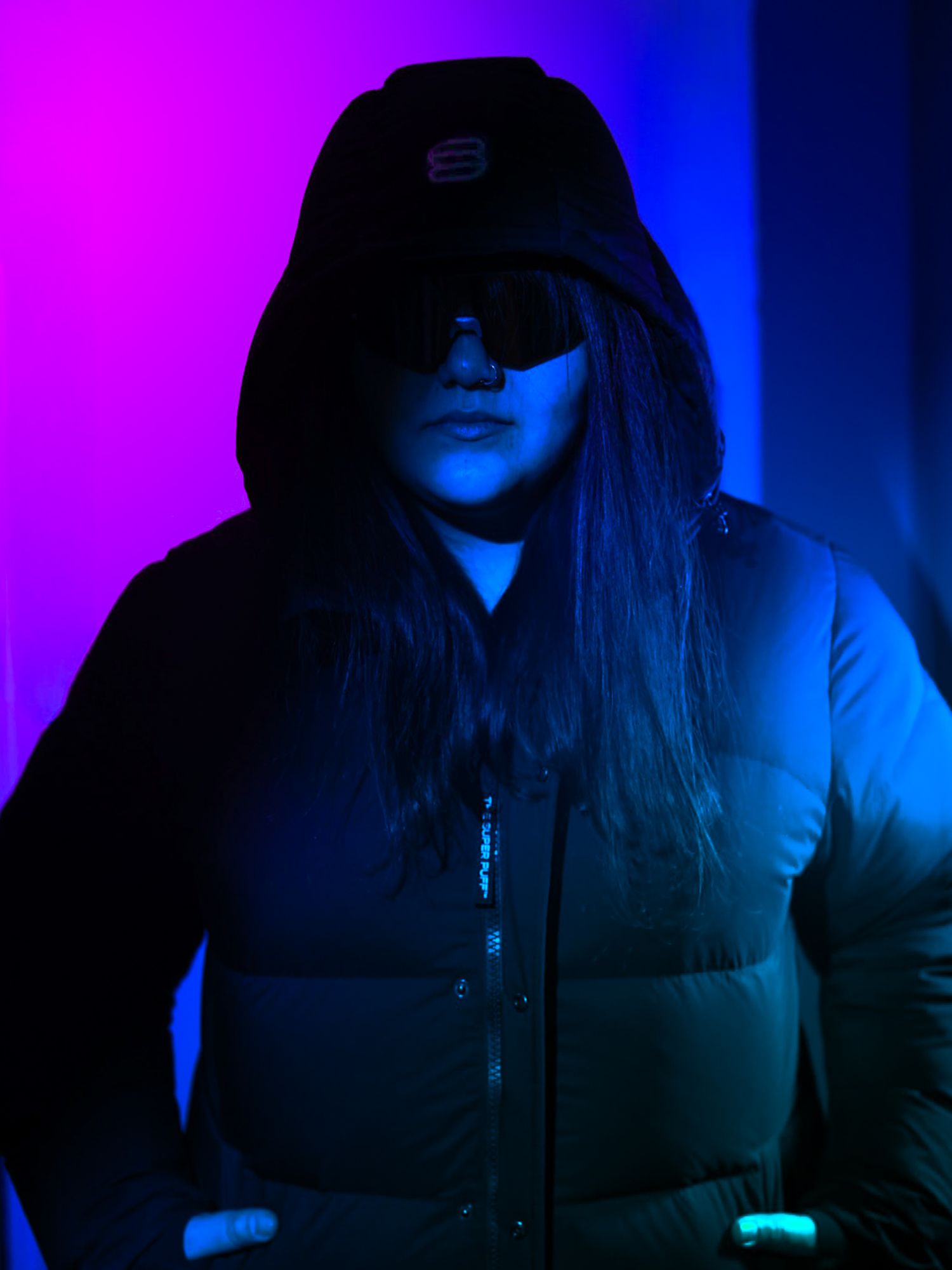 Figure in black hooded jacket and sunglasses posed against vivid purple and blue neon backdrop.