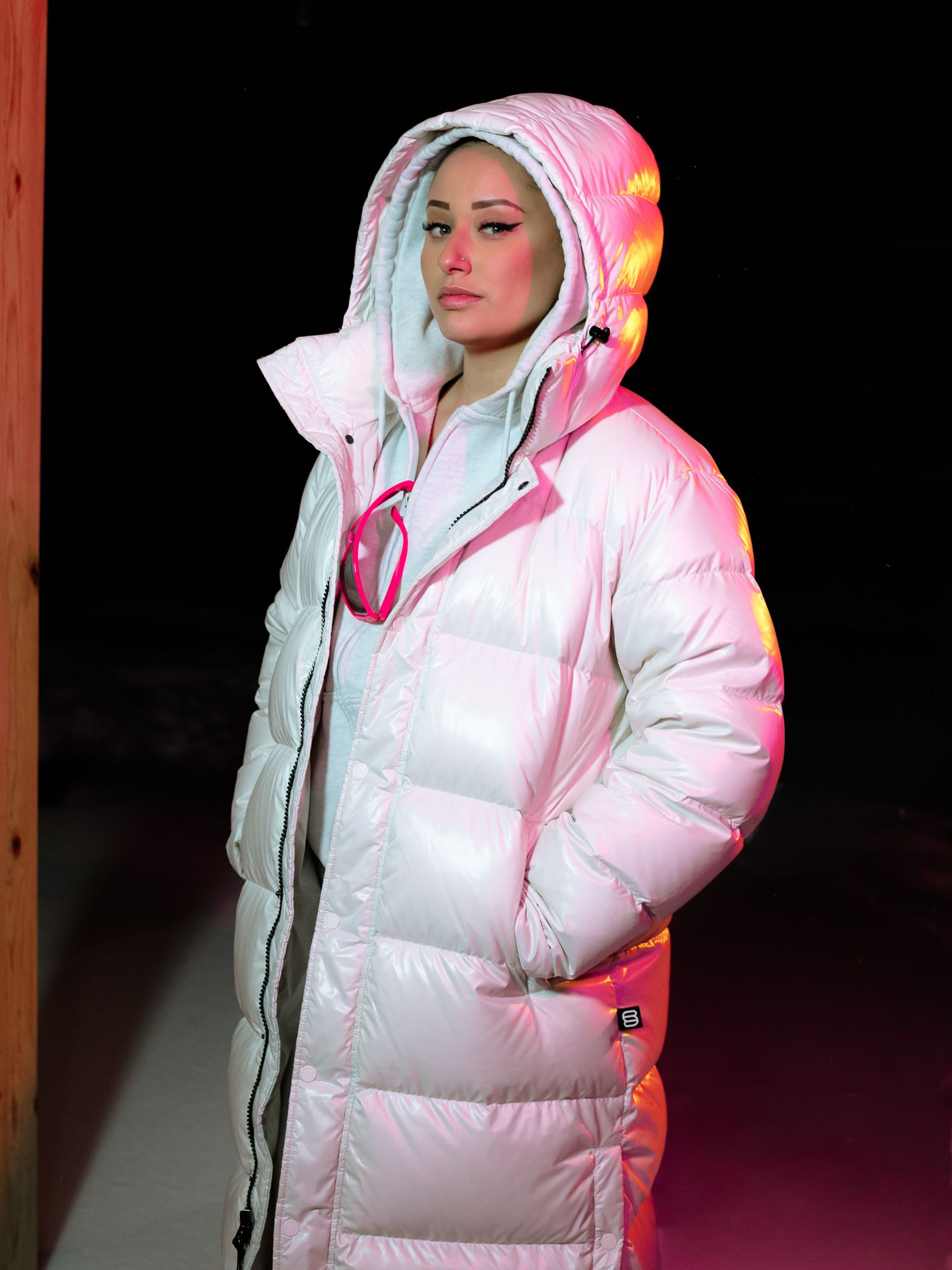 Woman wearing long white iridescent hooded puffer coat with pink sunglasses under dramatic pink lighting.