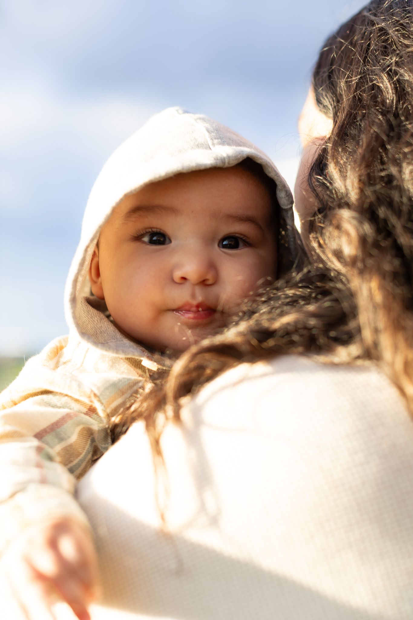 A baby wearing a white hat is held close in a loving embrace during an outdoor photoshoot.