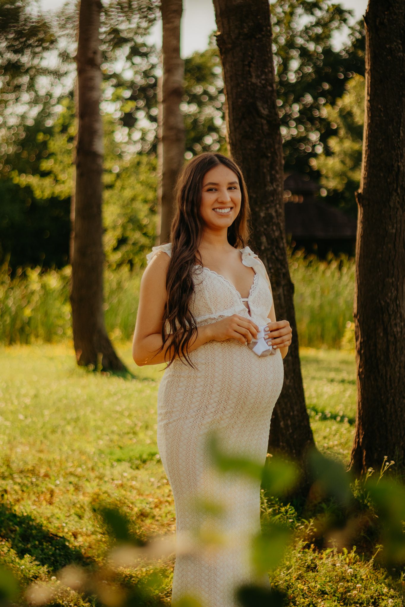 Maternity photo of woman in white dress standing among trees at golden hour.