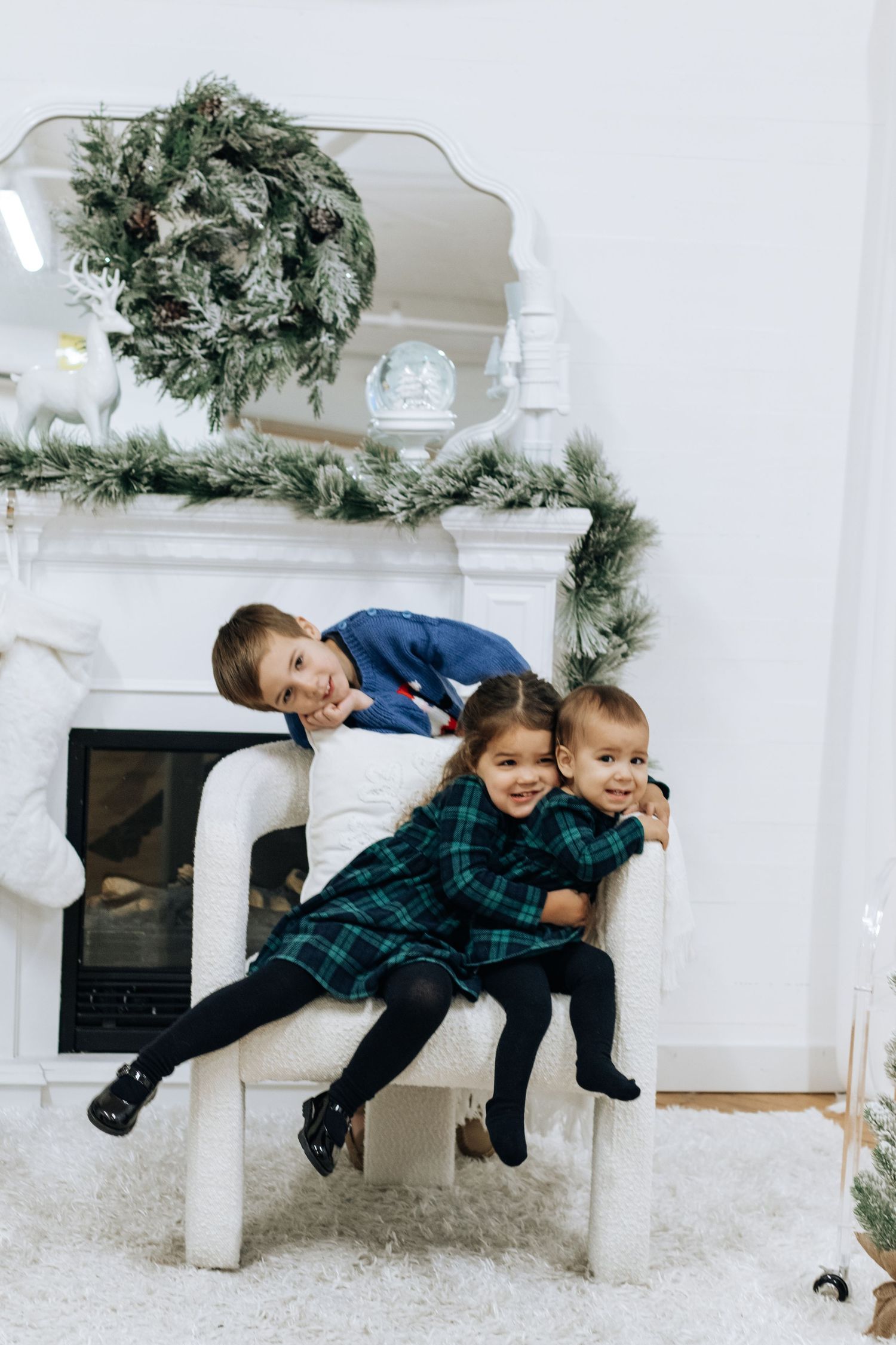 Children posing playfully on white bench with Christmas decorations and fireplace in background.