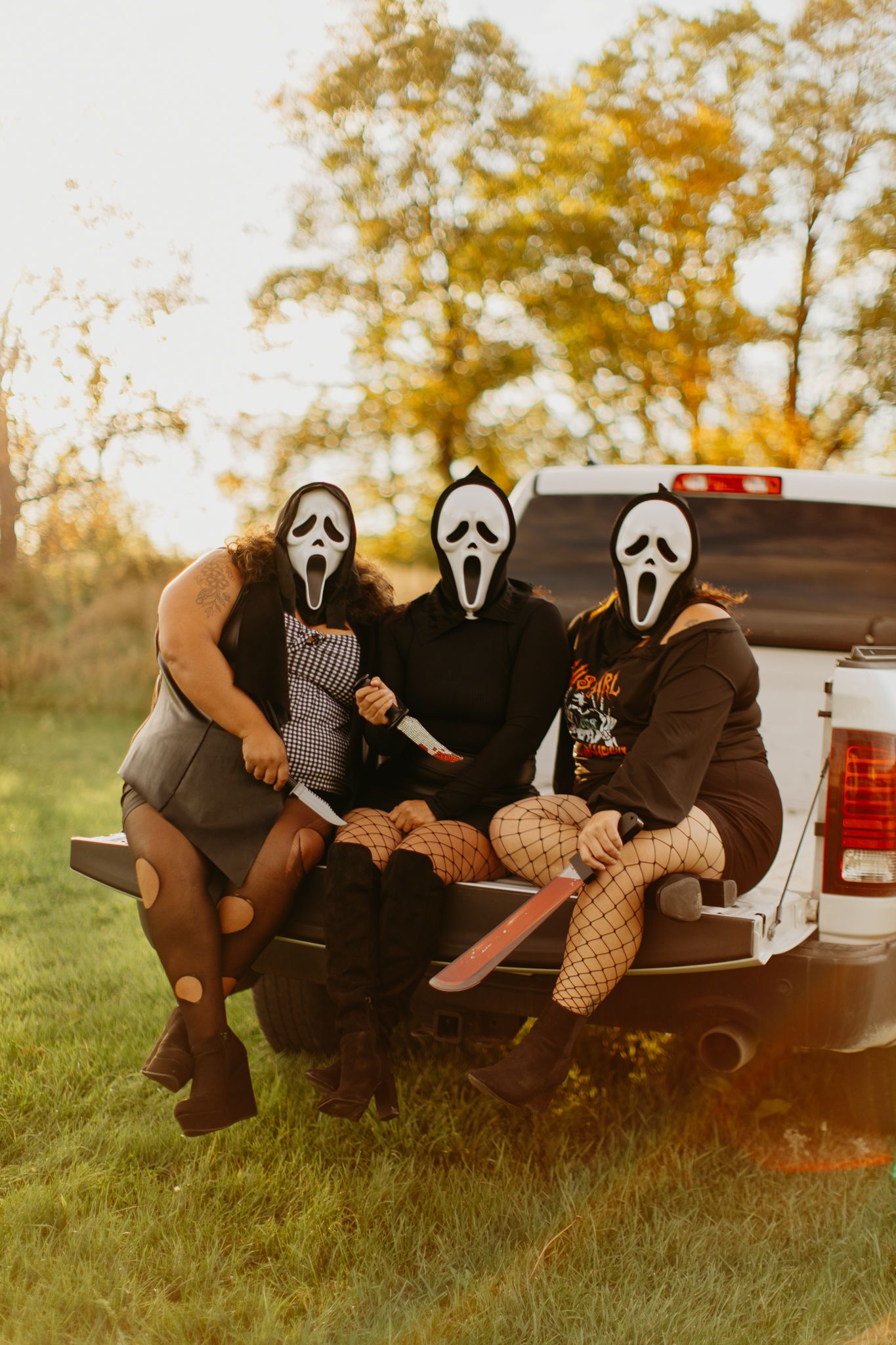 Three people wearing Scream masks sit on the tailgate of a pickup truck at sunset.