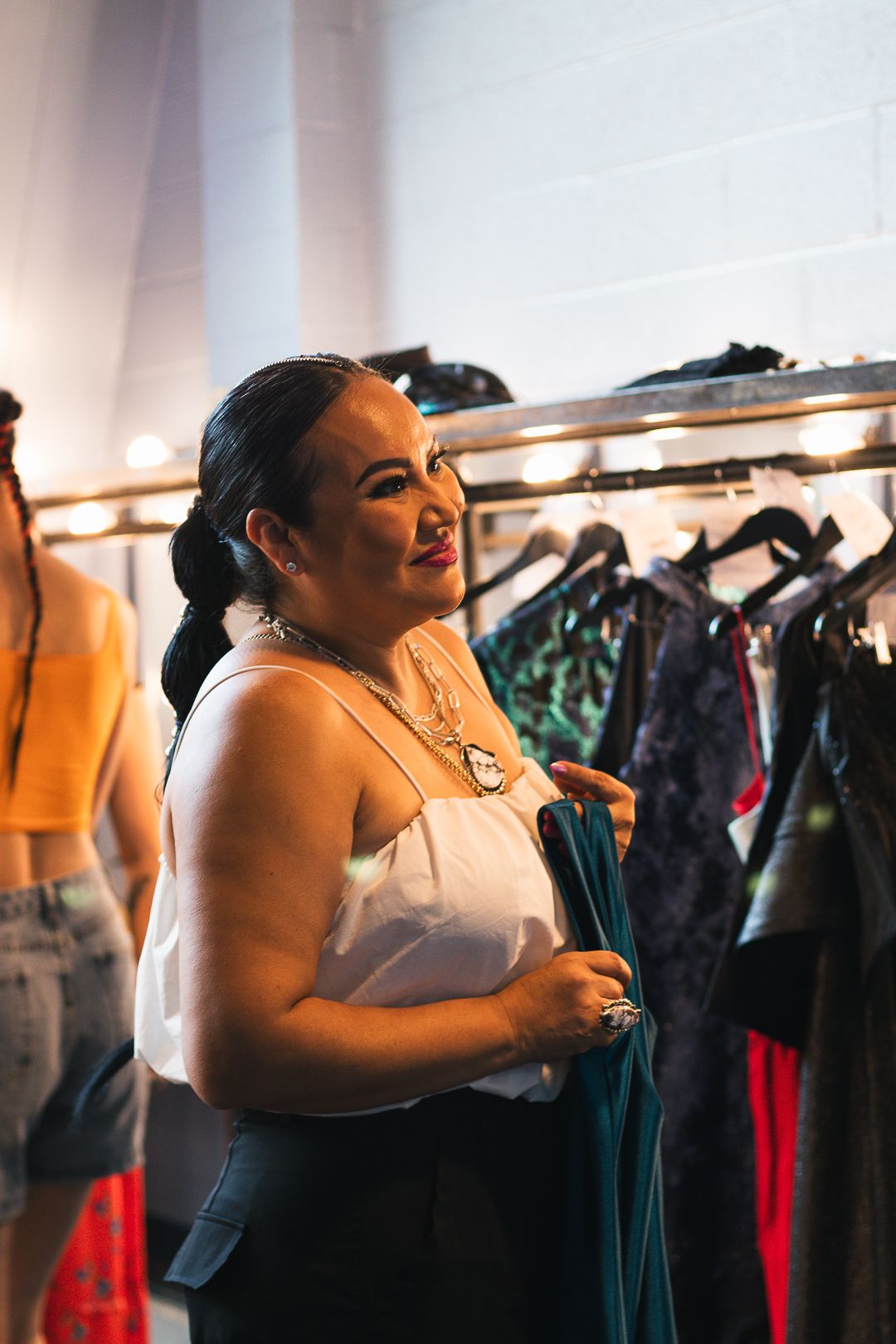 Woman smiles while browsing clothing rack backstage at fashion show wearing white top and silver necklace.