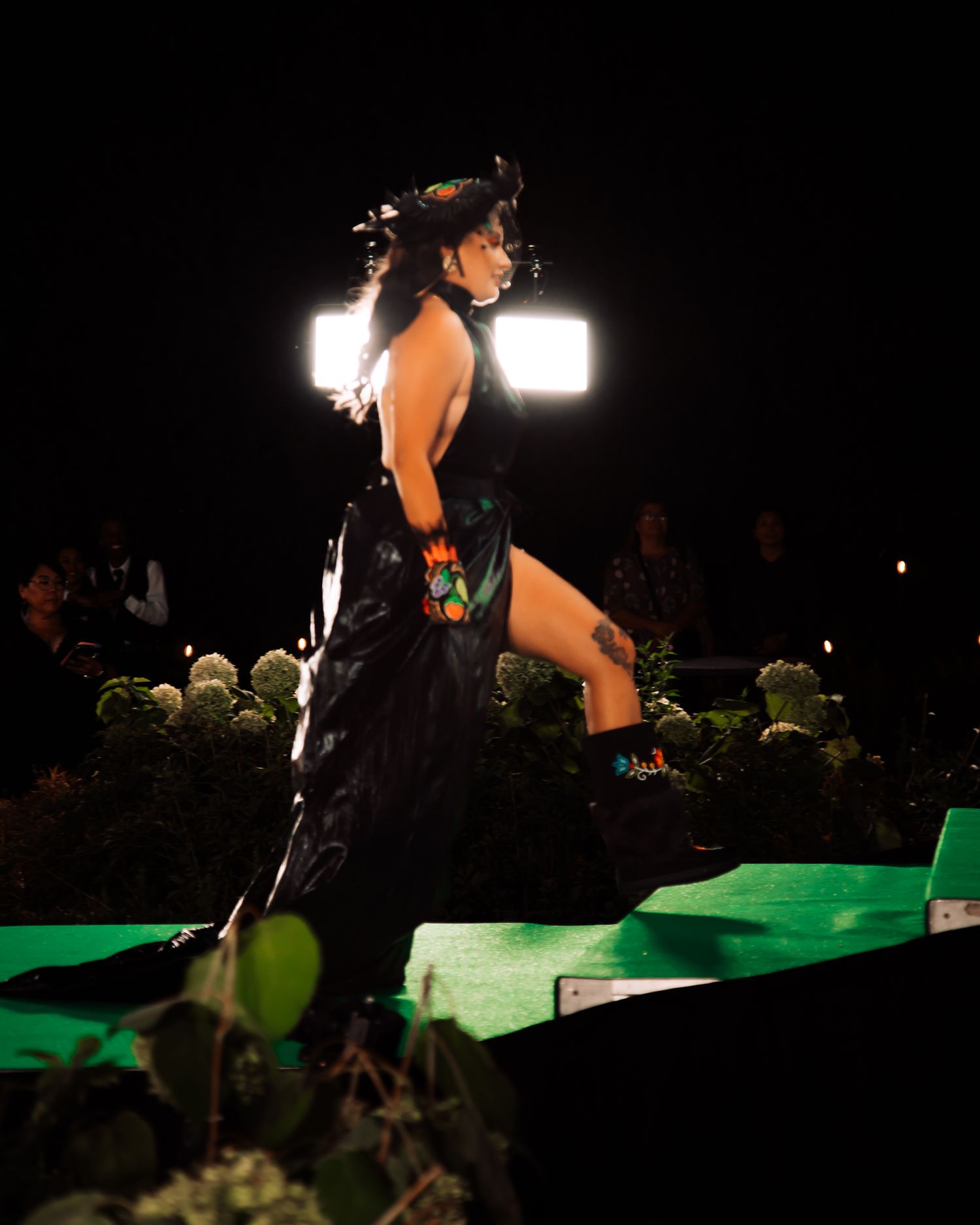 Model walking runway in dramatic black backless gown with floral embroidery at night.