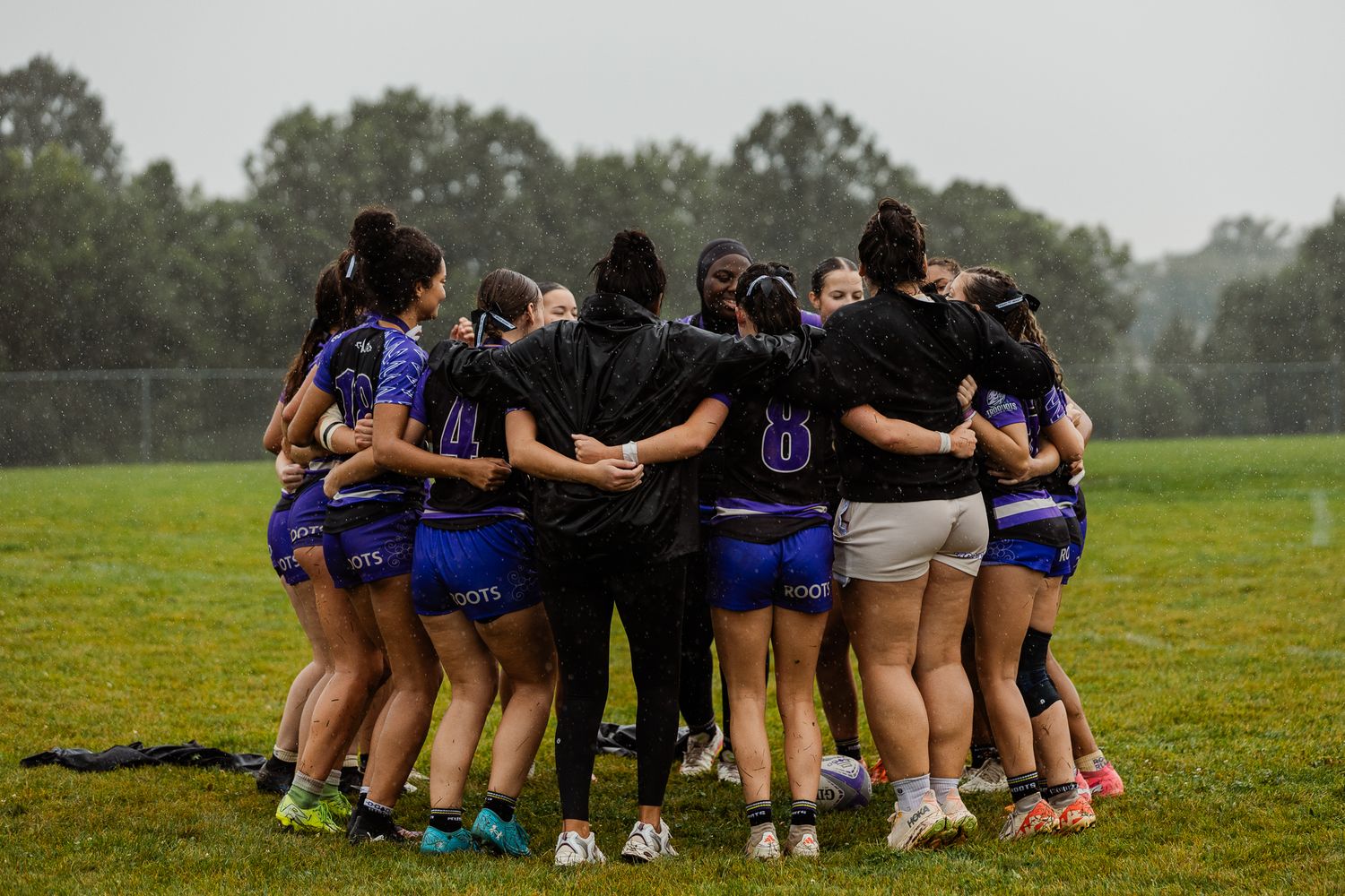 Girls rugby team huddles together in the rain on a green field during a wet weather match.