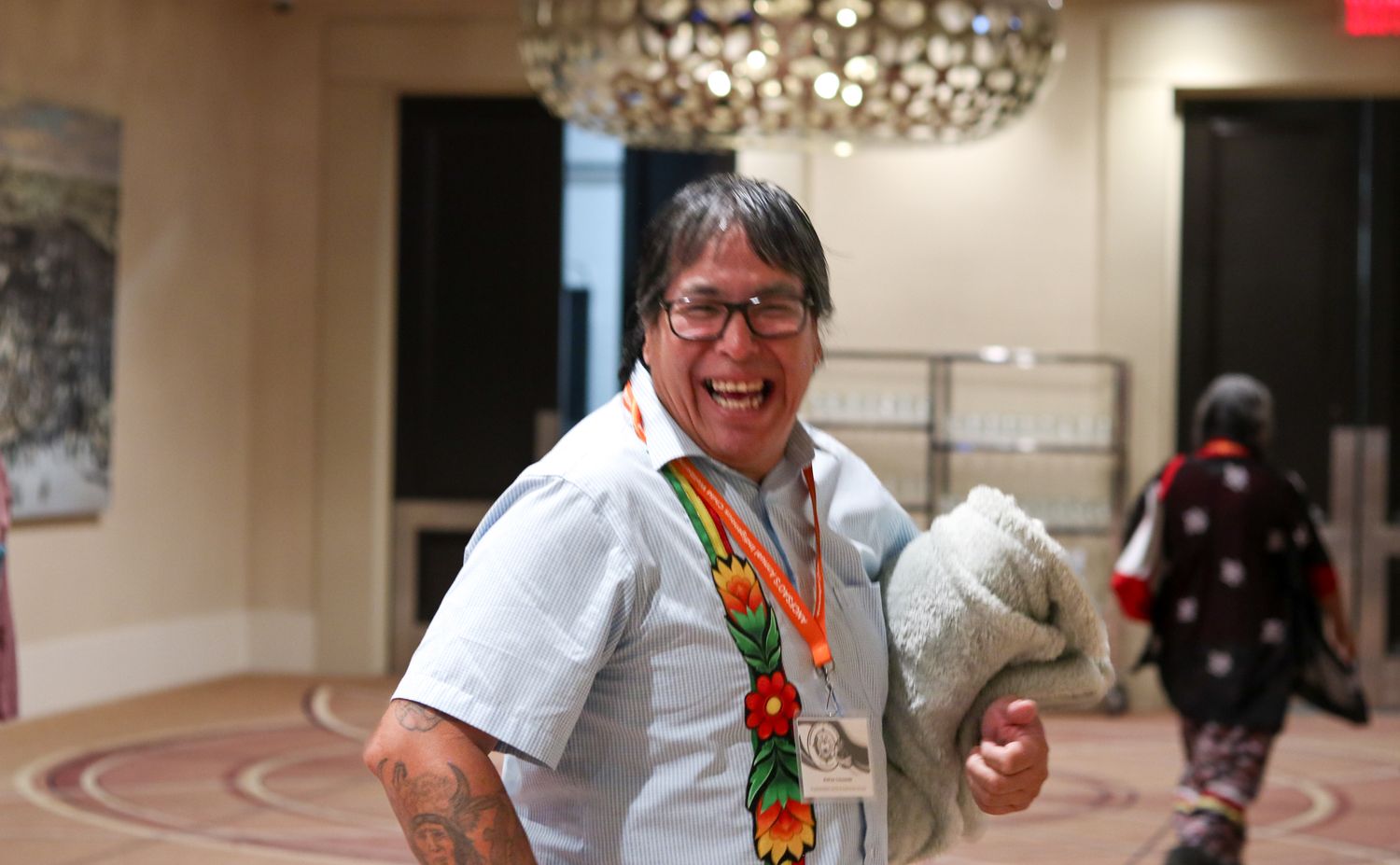 Smiling Indigenous person wearing traditional beaded medallion and white shirt holds stuffed animal in elegant venue.