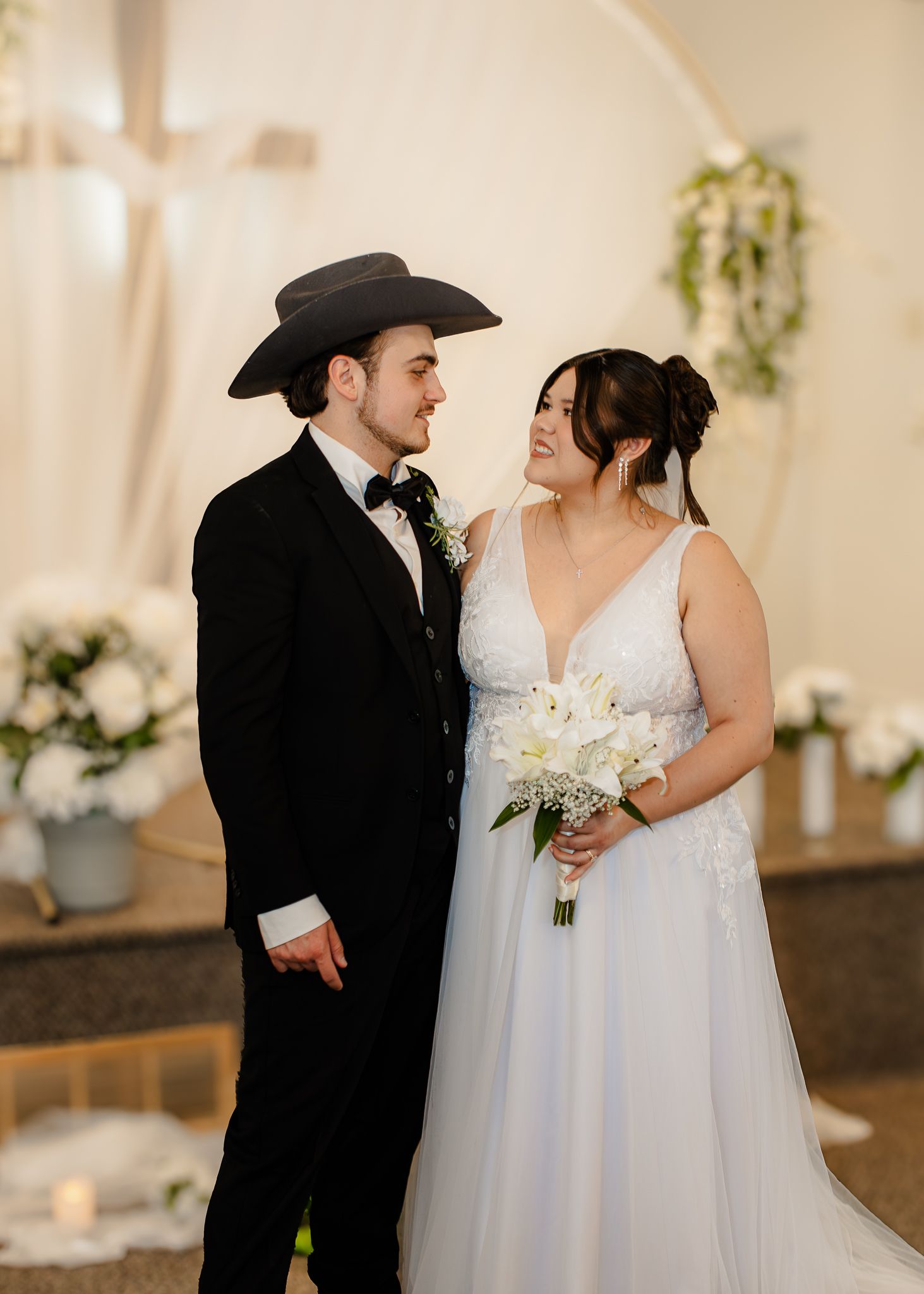 Bride in white gown and groom in black tuxedo with cowboy hat smile at each other at wedding altar.