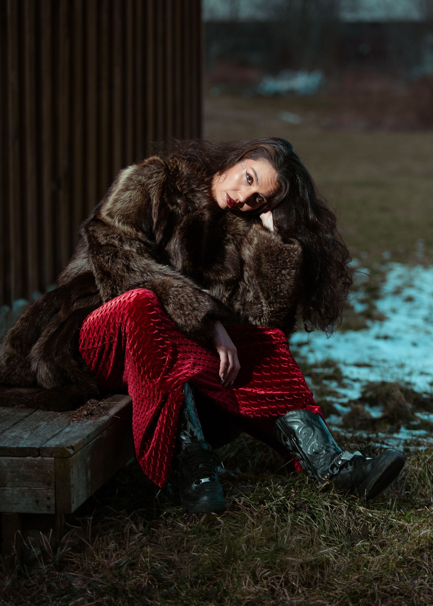 Woman in brown fur coat and red velvet pants sitting outdoors on wooden bench in winter.