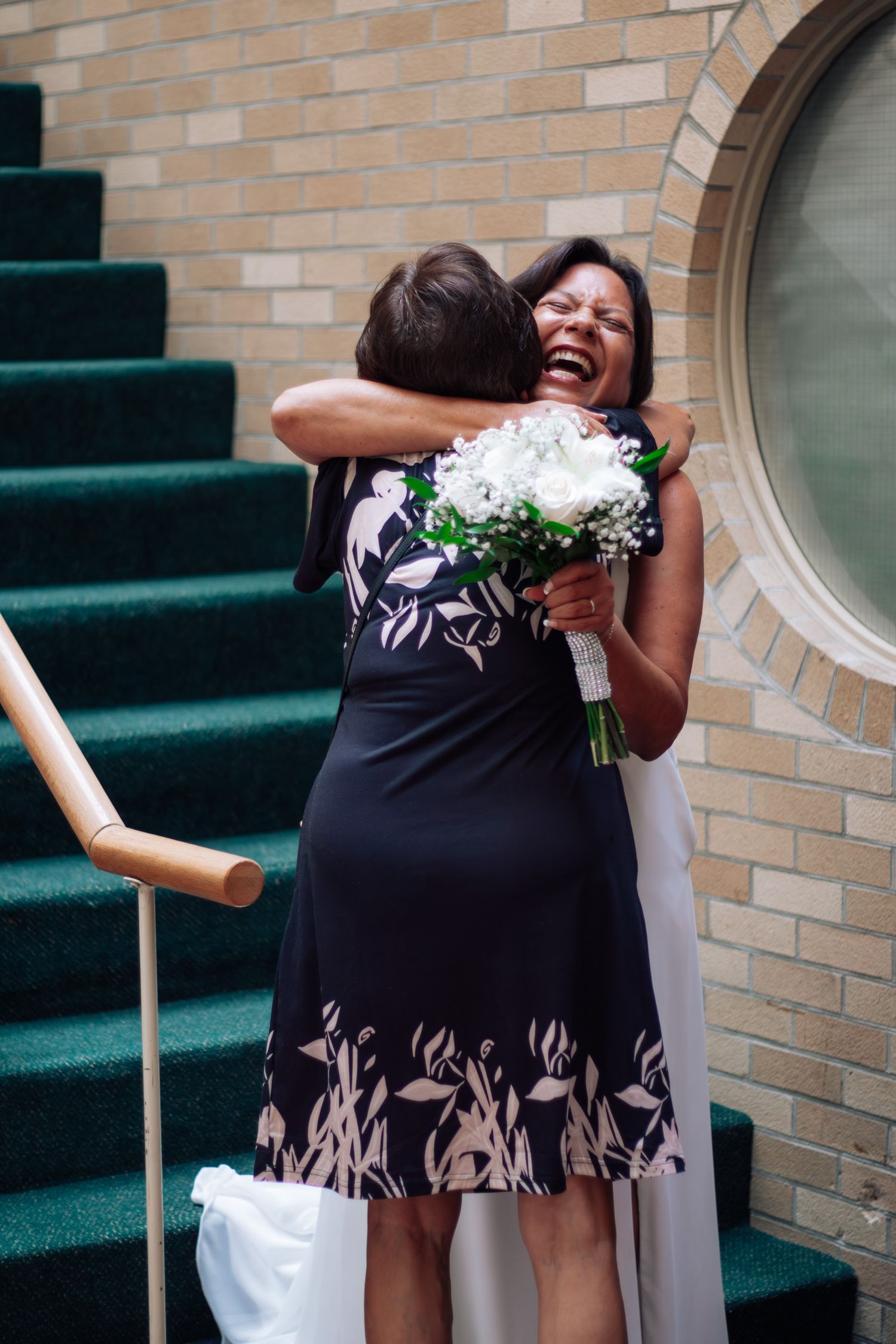 Joyful bride holding white bouquet laughing while embracing guest on green carpeted church steps.