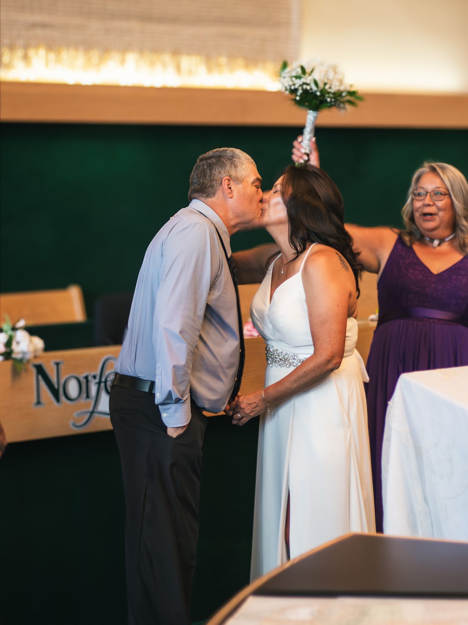 Bride and groom share first kiss at wedding ceremony while bridesmaid in purple dress looks on.