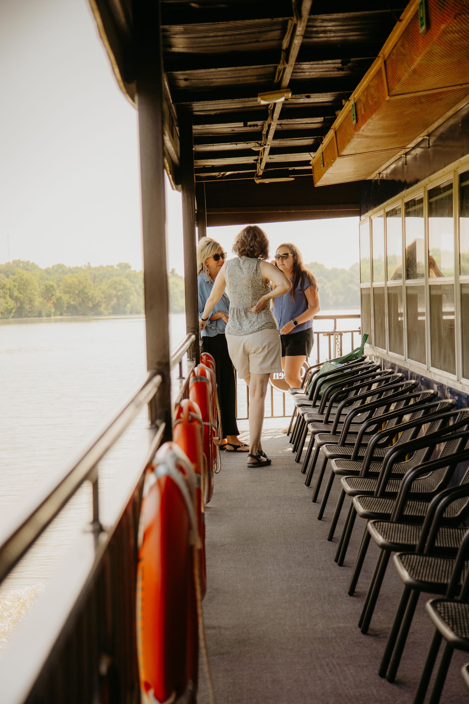 Empty rows of black chairs line the deck of a riverboat overlooking the water.