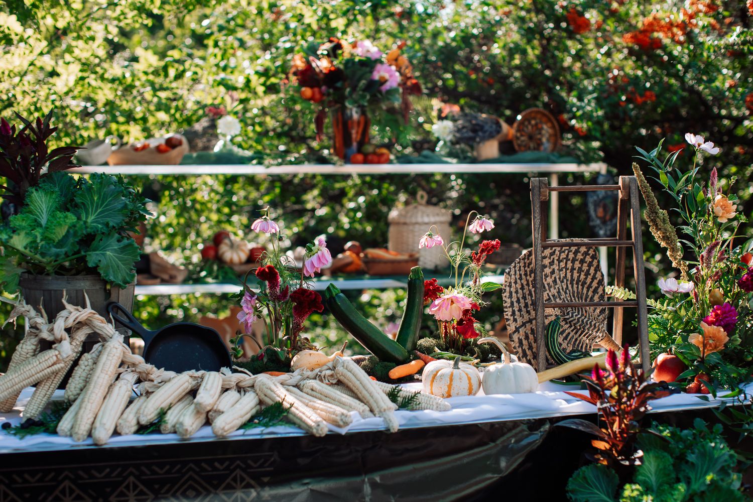 Outdoor garden buffet table with fresh corn and vegetables, surrounded by lush greenery and colorful flowers.