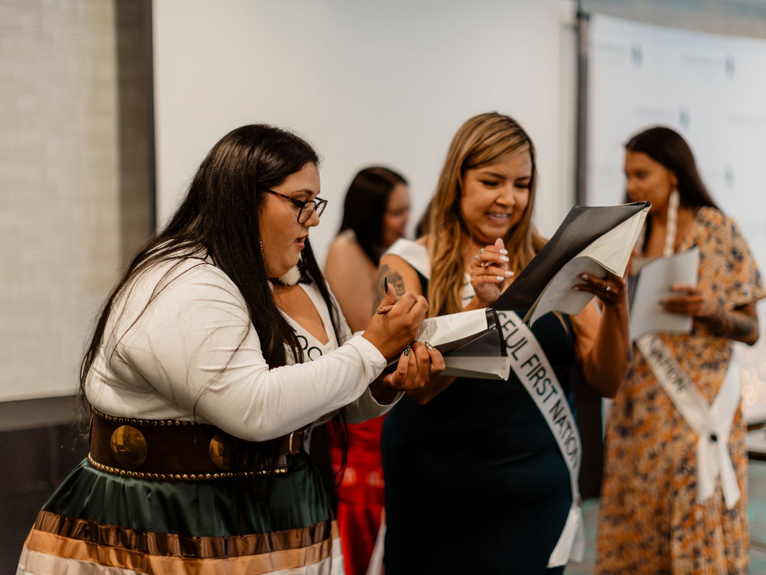 Two women in traditional Indigenous regalia examine a sash at a First Nations pageant ceremony indoors.