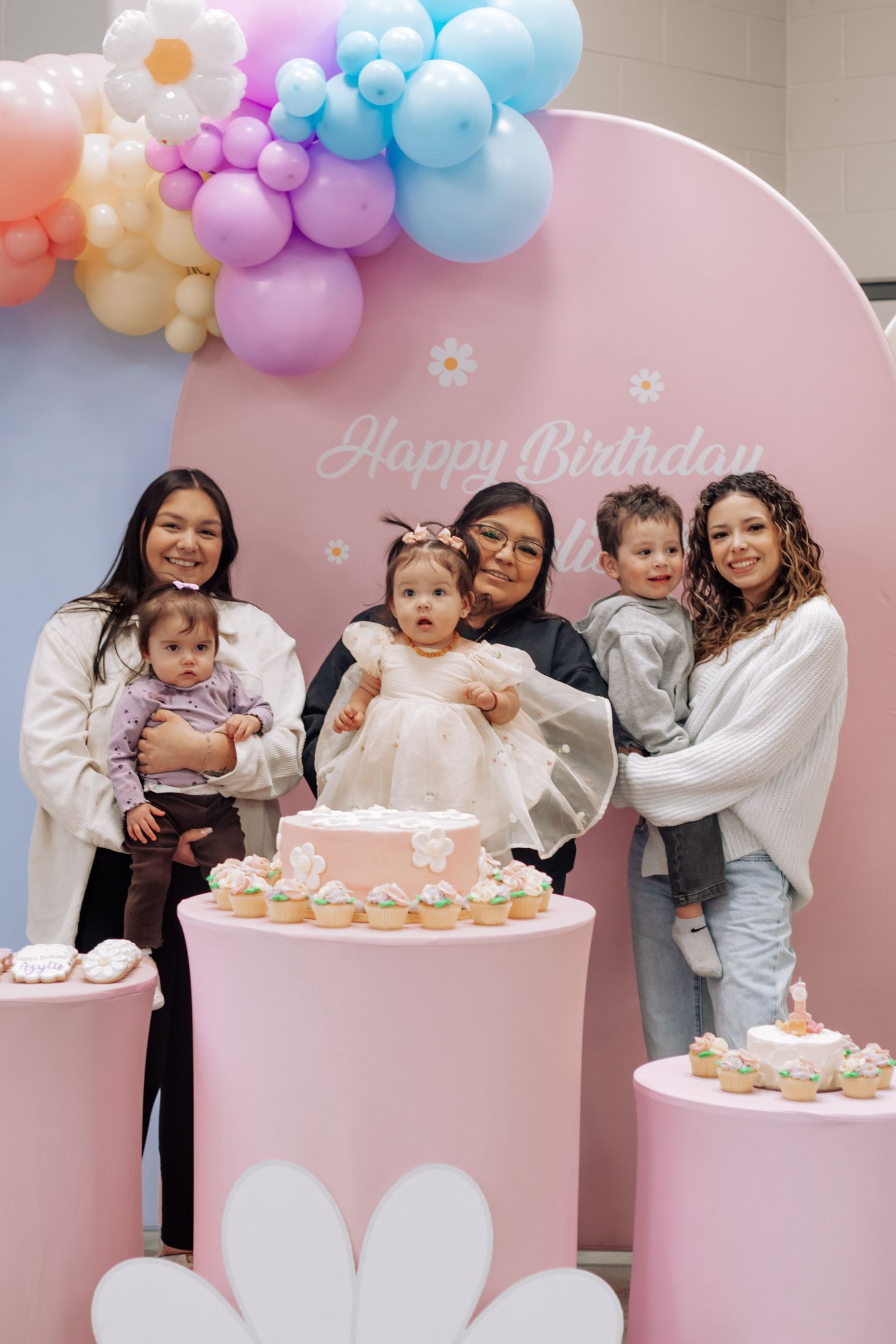 Three women holding babies pose by a pink birthday cake and balloon arch at a colorful first birthday party celebration.