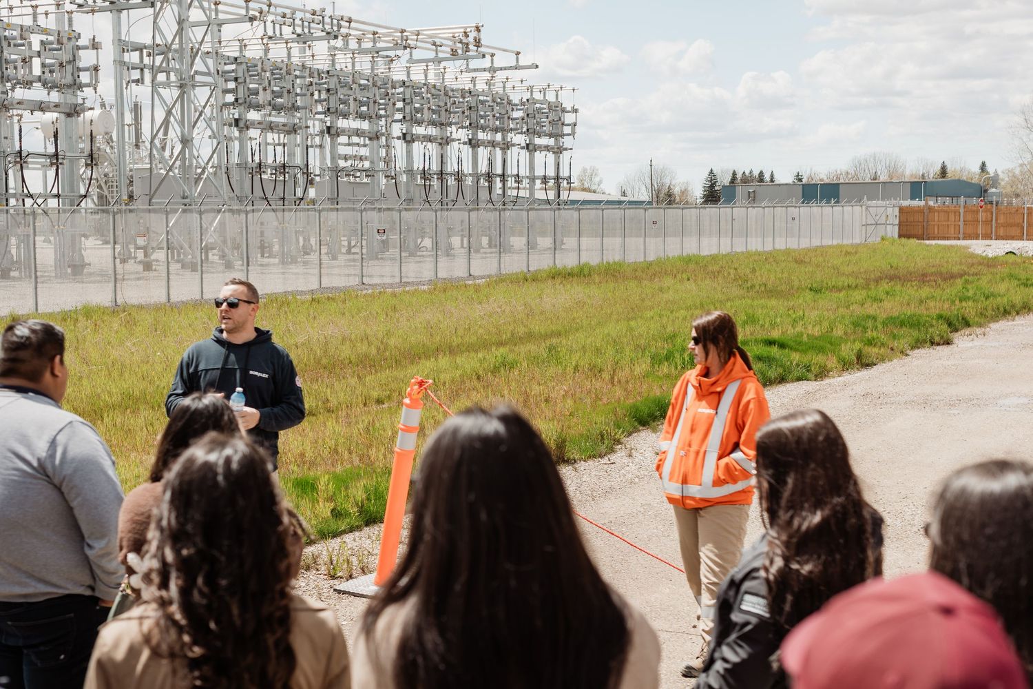 Two presenters in safety gear address a group of visitors outdoors near an electrical substation facility.