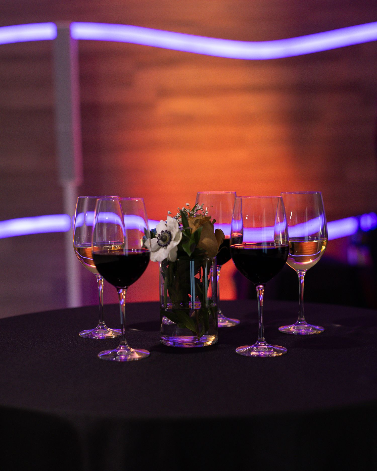Elegant wine glasses filled with red wine arranged on a dark tablecloth with a floral centerpiece under warm ambient lighting.