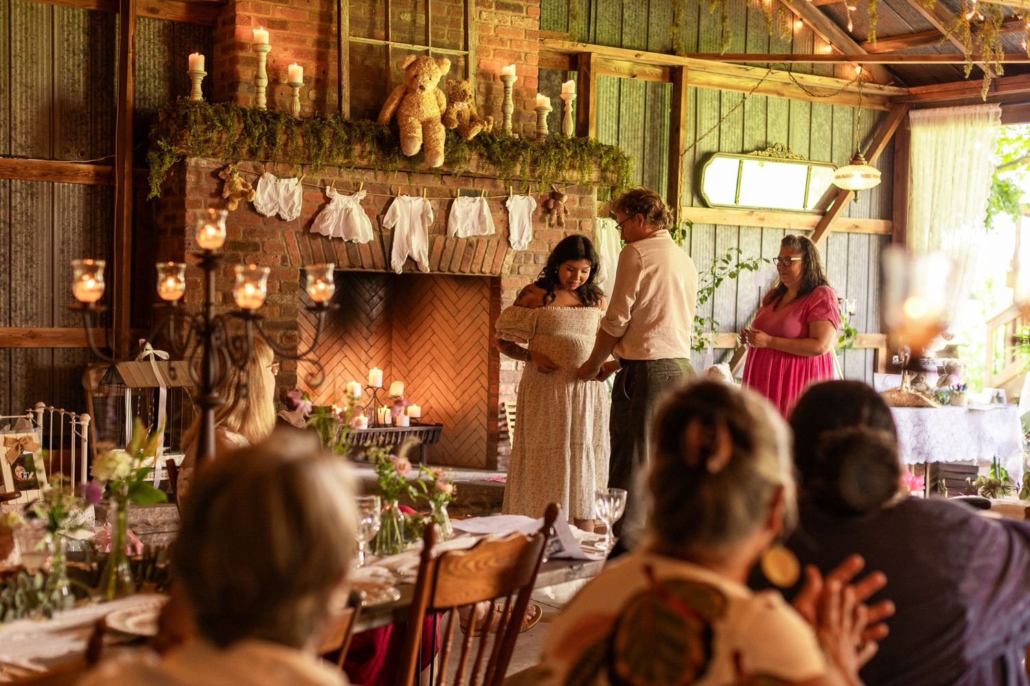 Rustic barn baby shower with couple opening gifts near candlelit fireplace decorated with baby clothes and greenery.