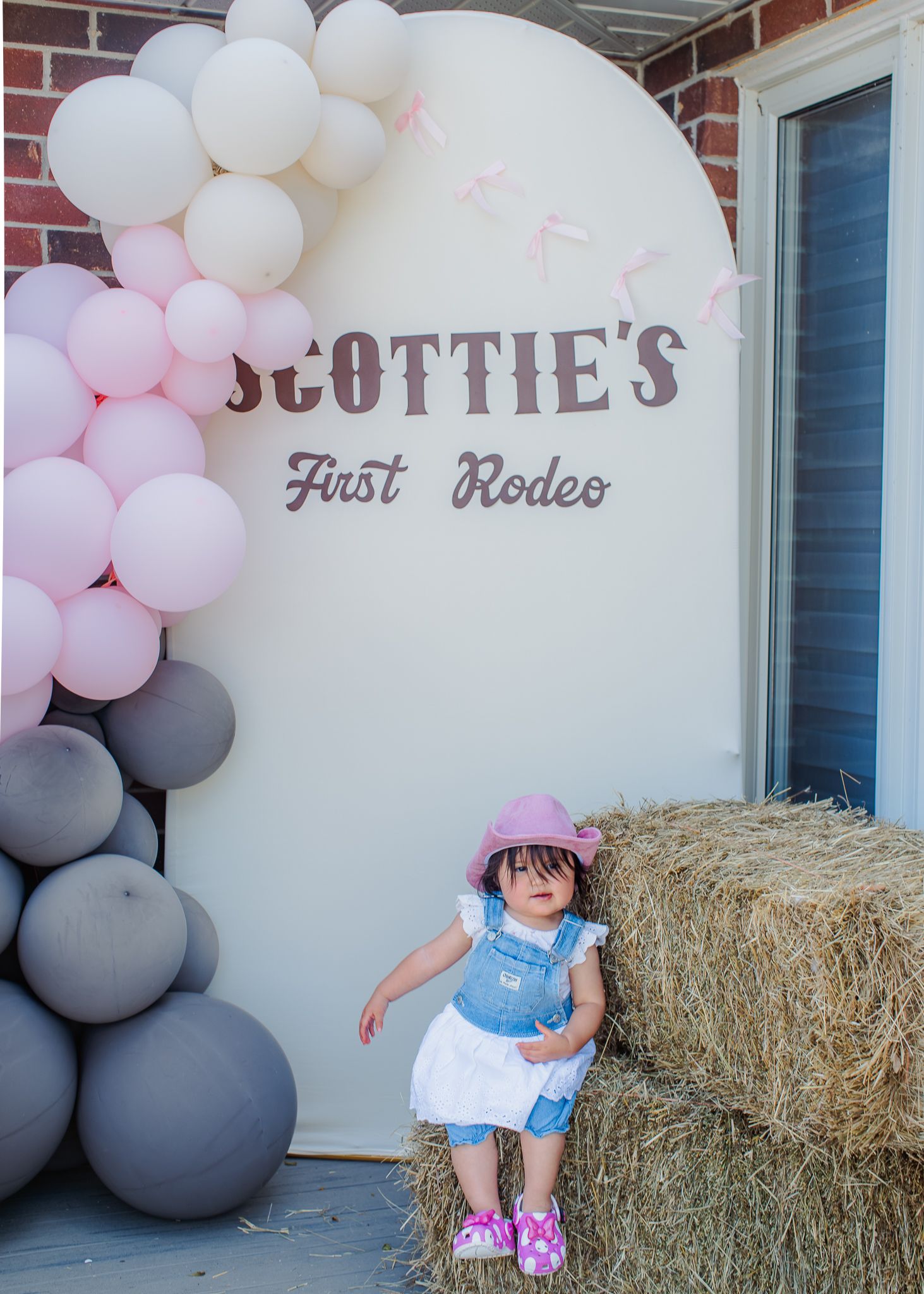 Toddler in pink cowboy hat and denim overalls poses by hay bale at Scottie's First Rodeo birthday party.