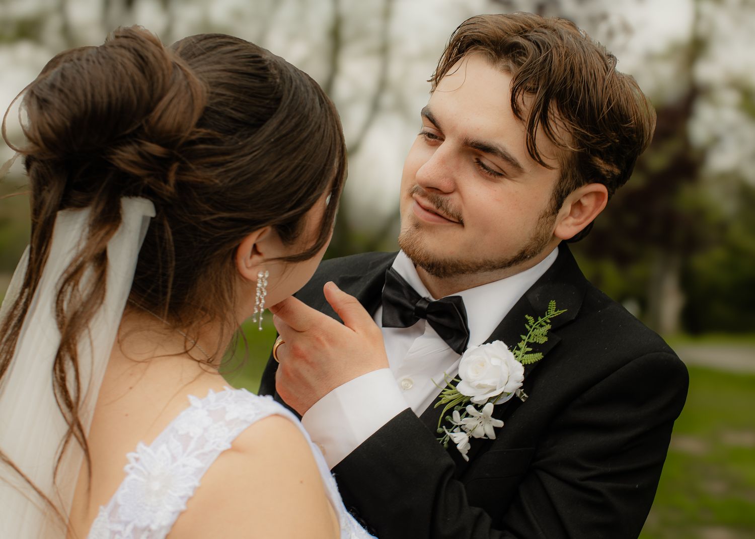 Groom in black tuxedo smiles tenderly at bride in white dress during outdoor wedding ceremony.