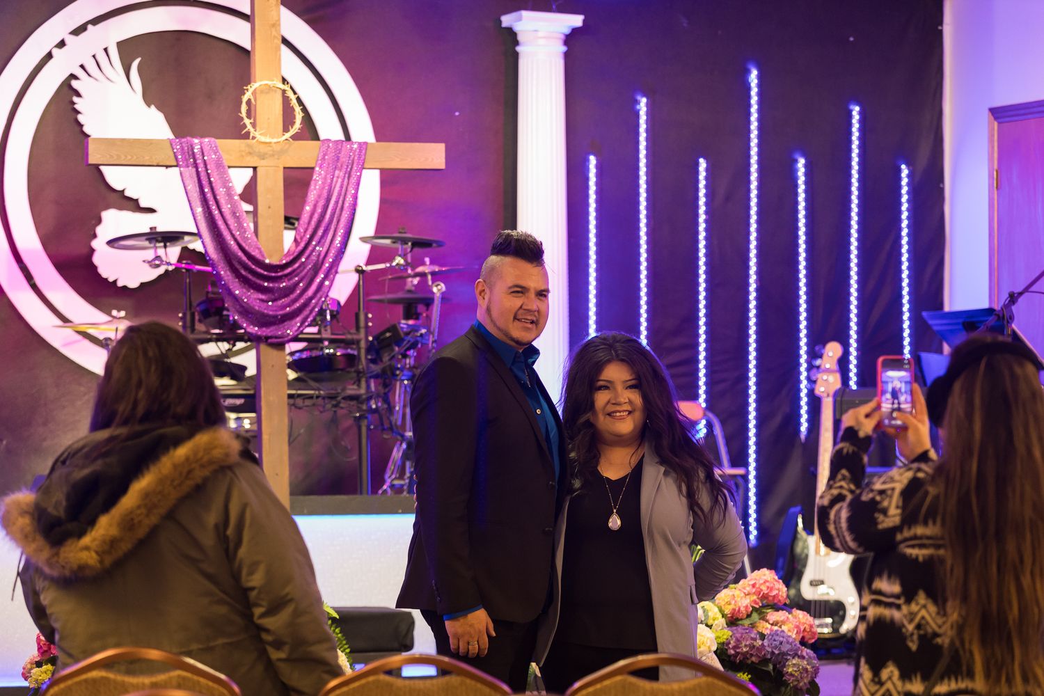 A man and woman pose together inside a church with a cross and purple decorations in the background.