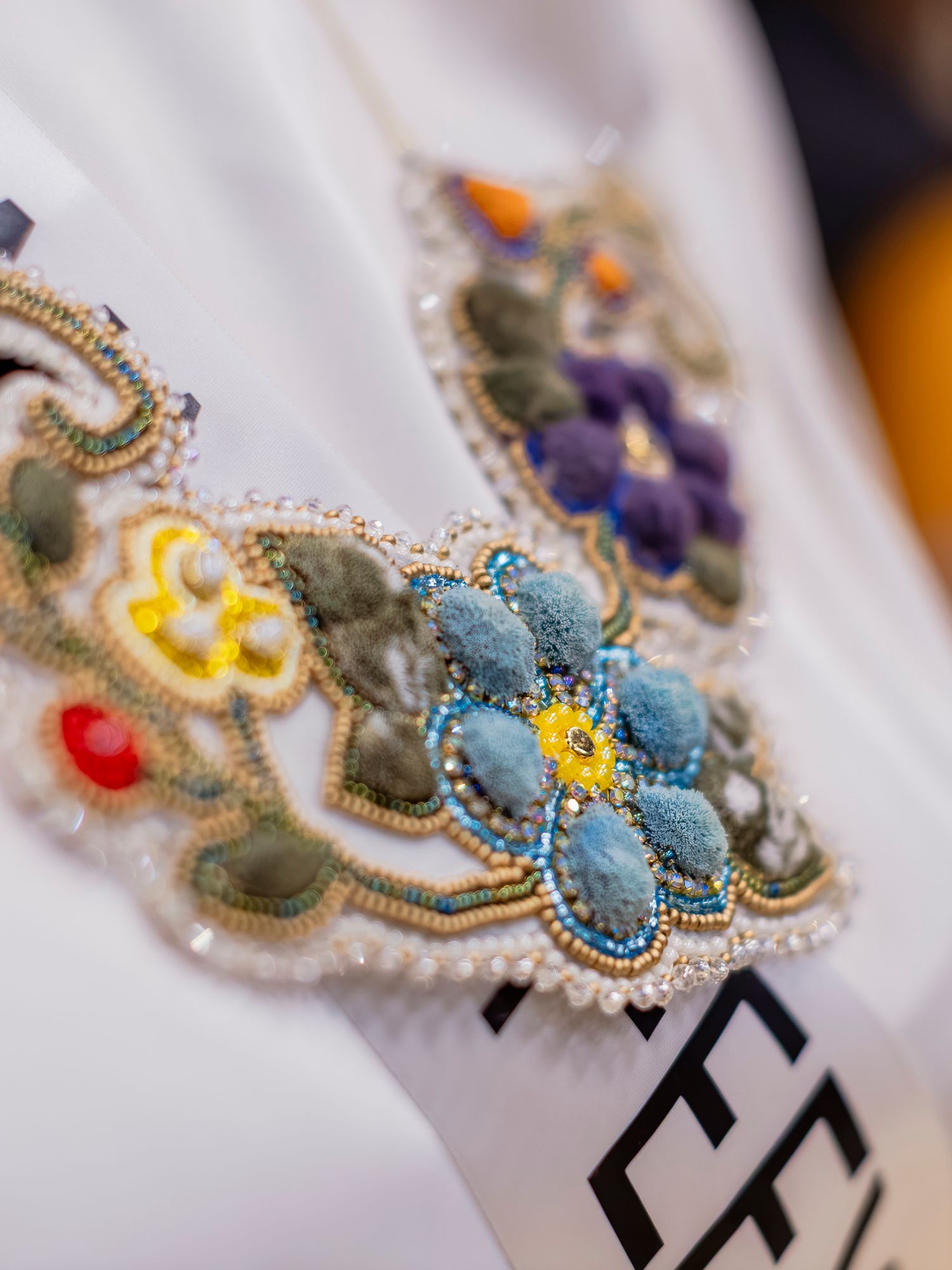 Intricate colorful beaded embroidery necklace with floral patterns in blue, yellow, and purple on white fabric.