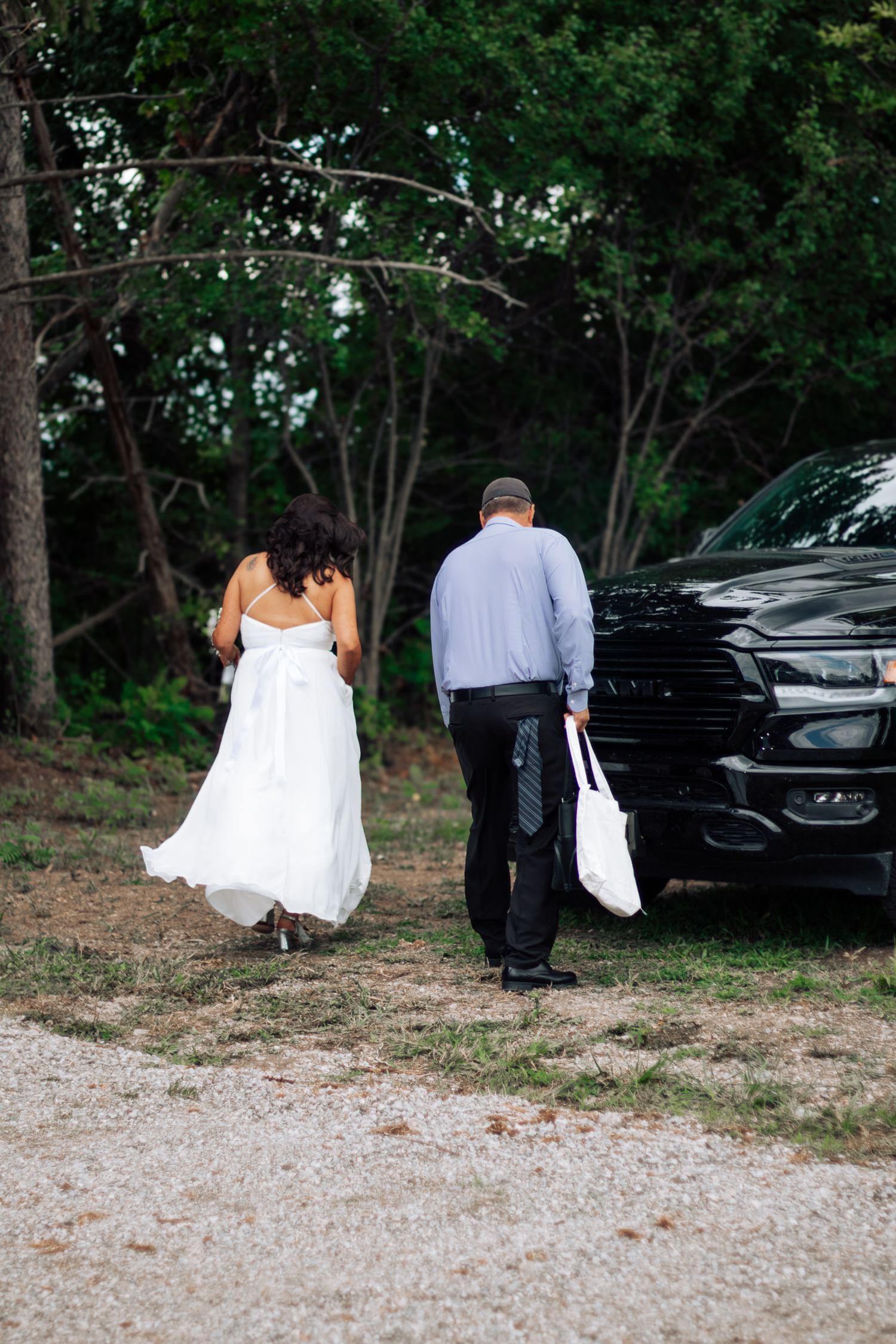 Bride in white gown and groom in blue shirt walk together toward a black SUV in a wooded outdoor setting.