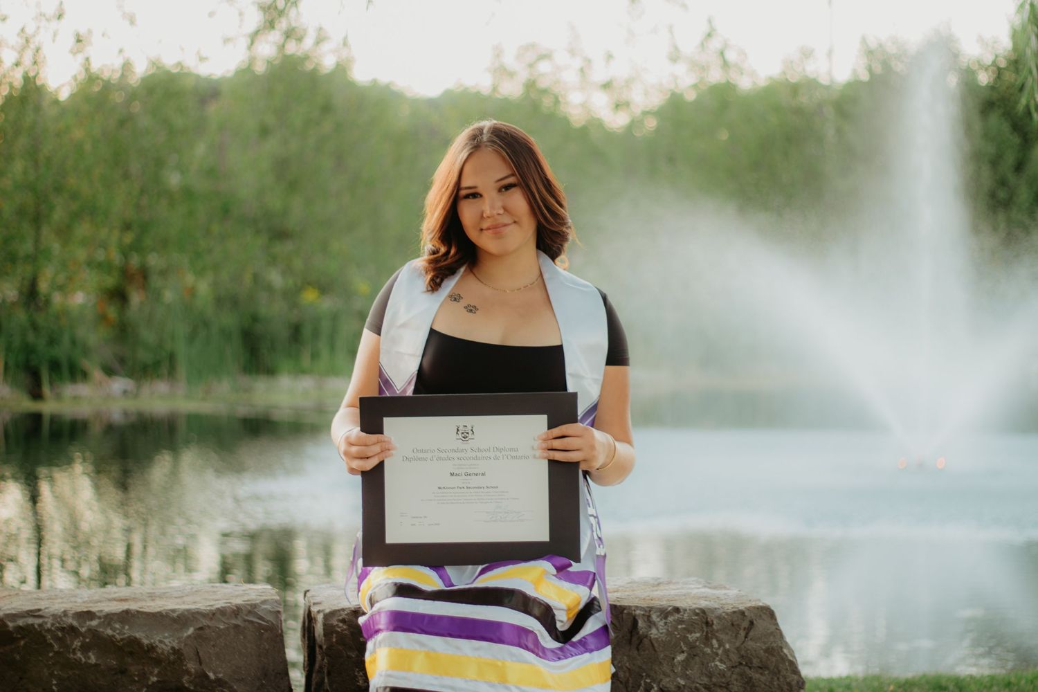 A graduate holds their diploma while posing for photos by a misty lake.