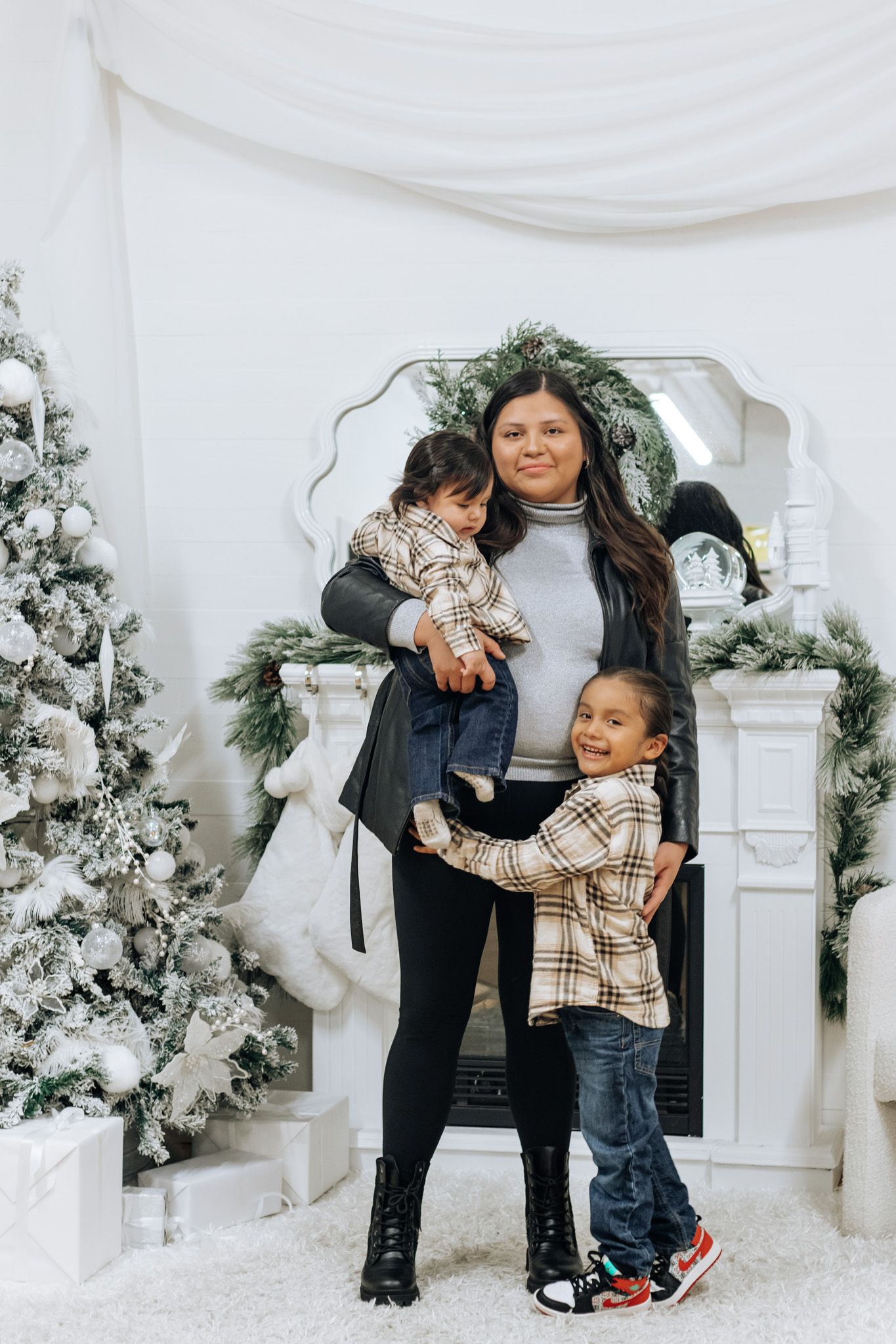 Heartwarming family portrait against white Christmas trees and decorated fireplace backdrop.