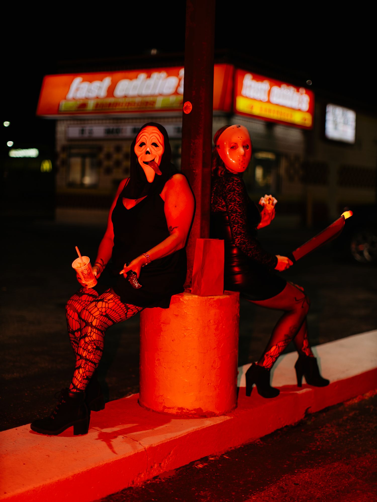 Red-tinted nighttime scene with figures near a cylindrical object outside a fast food restaurant.