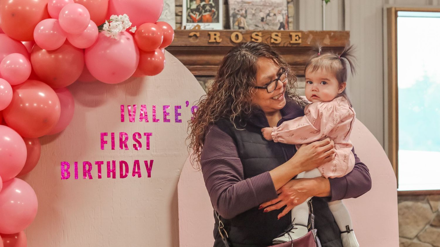 Woman holds baby at Ivalee's First Birthday party with pink balloon arch decoration.