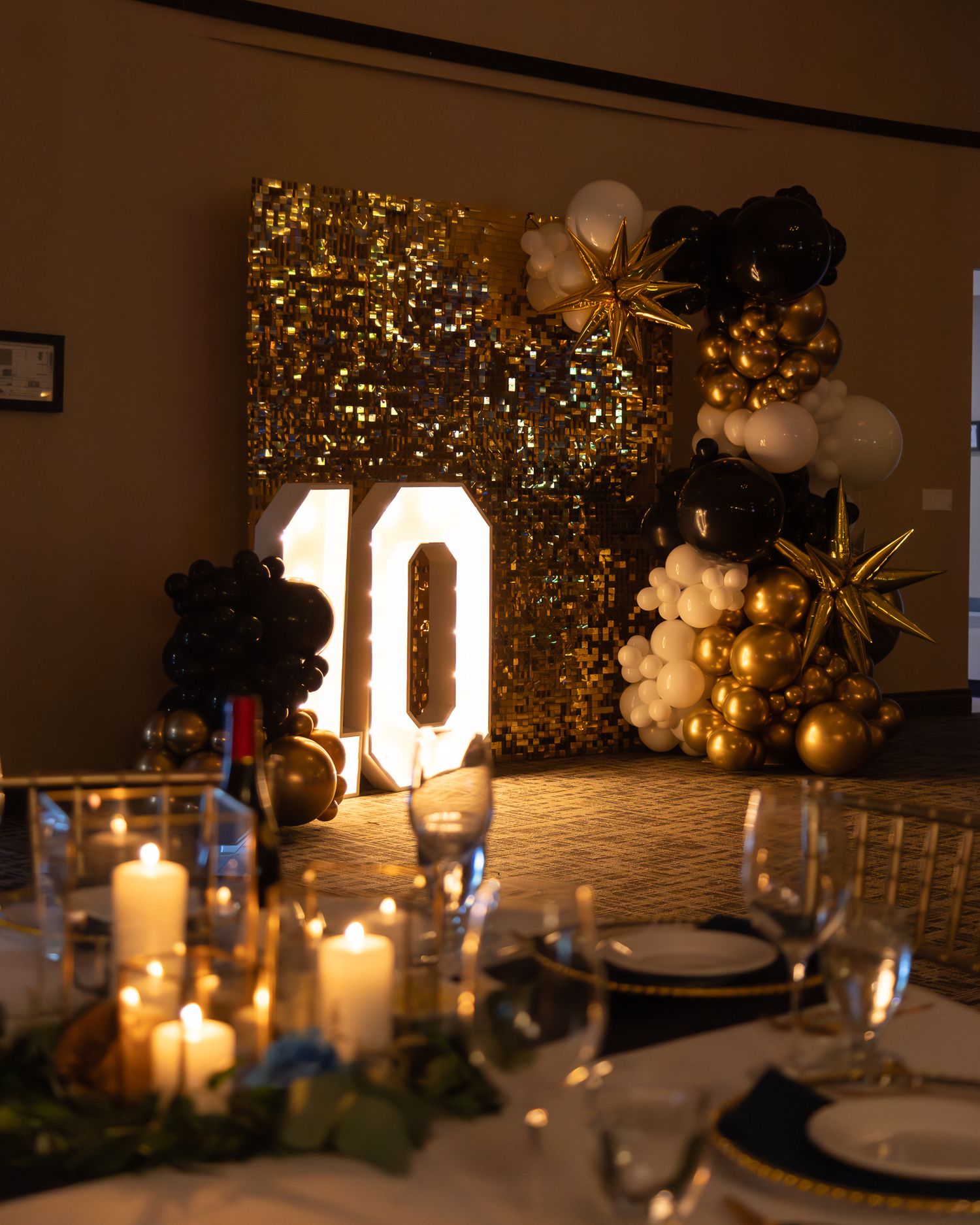 Gold sequin backdrop with illuminated number 10, black and gold balloon arch, and elegant candlelit table setting.