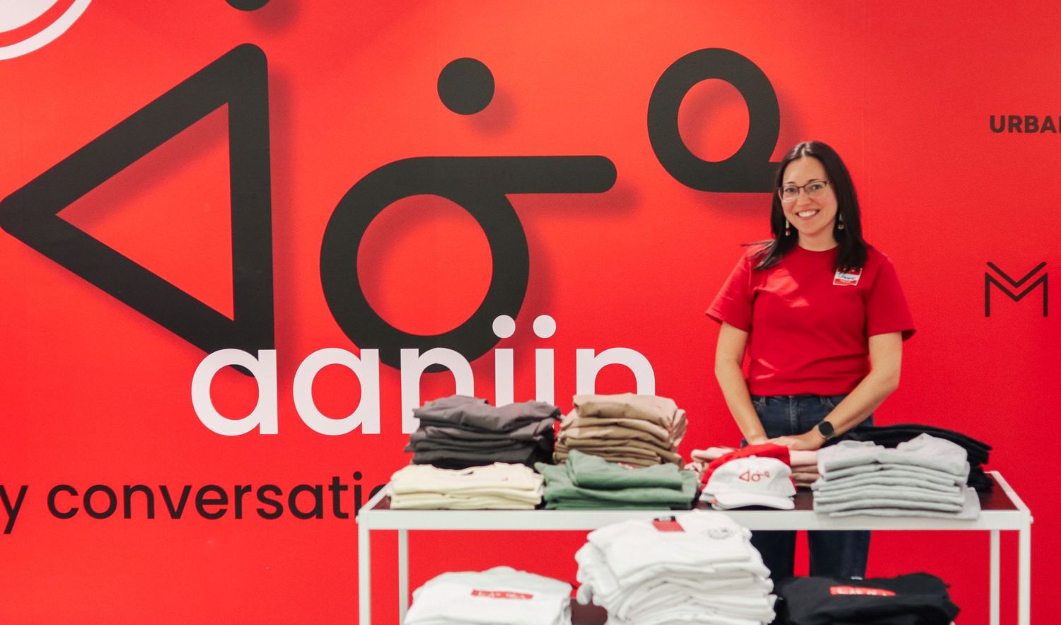 Woman in red shirt stands smiling beside a display table of folded shirts at the Aapiin conversational language booth.