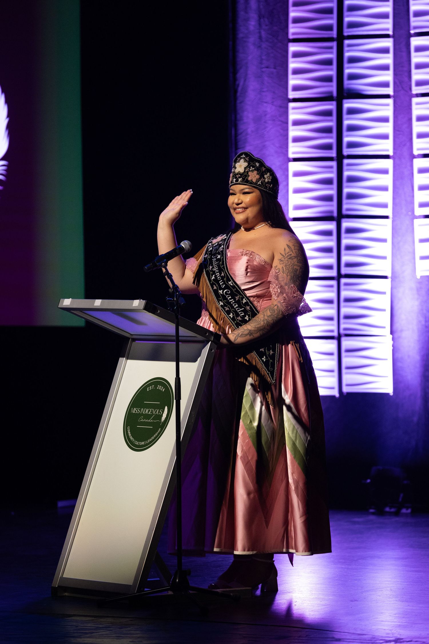 Woman in elegant pink gown and crown waves on stage at Miss Indigenous Canada pageant event.