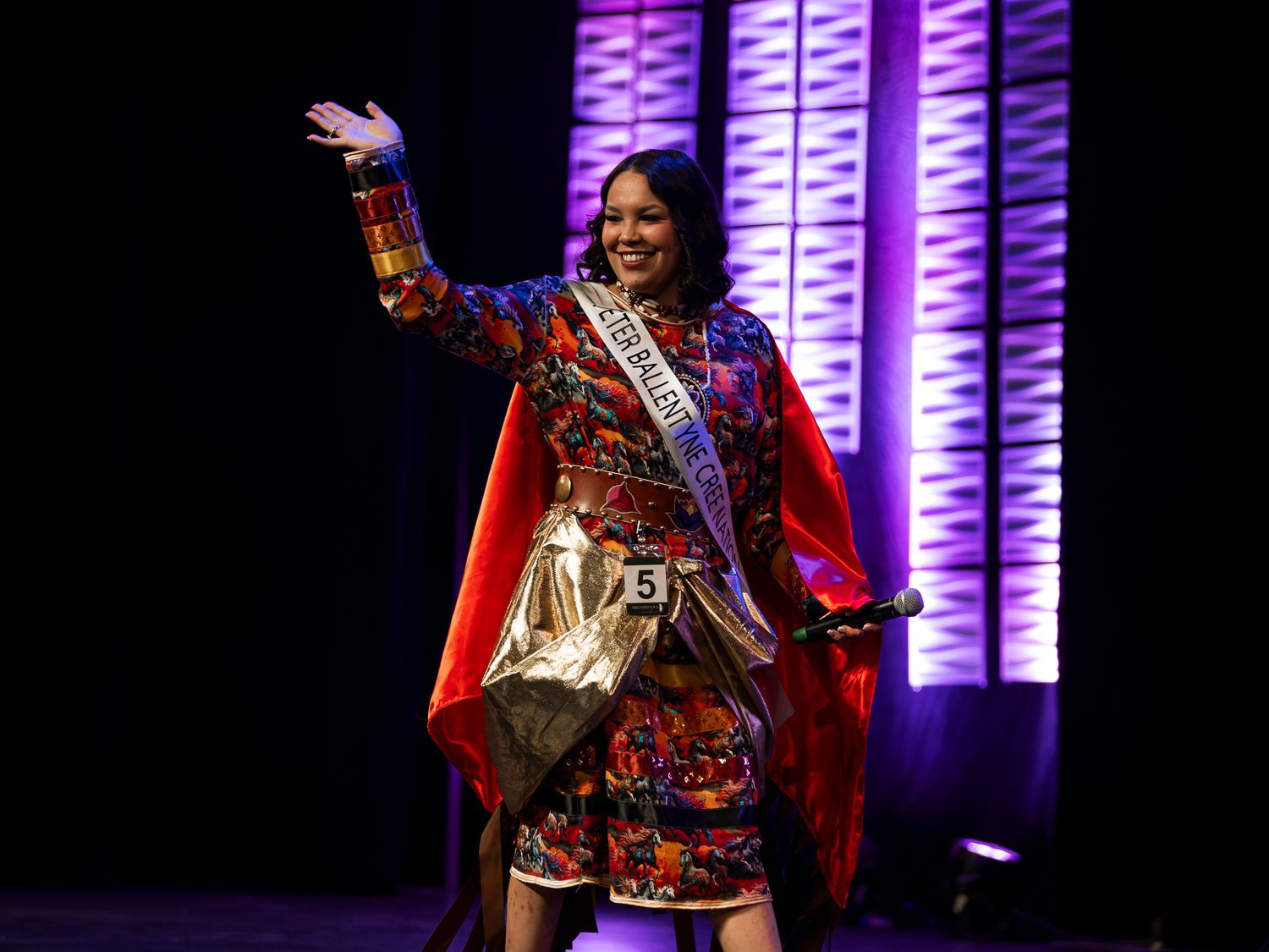A pageant contestant in colorful Indigenous regalia waves to the crowd on a purple-lit stage.