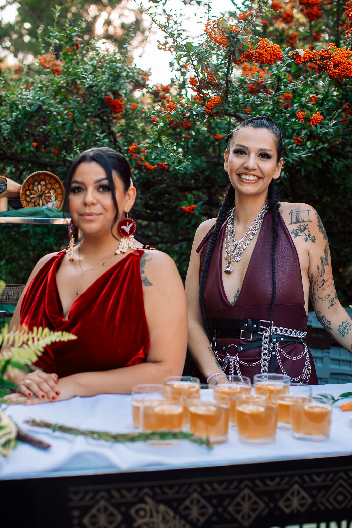 Two tattooed women in red and burgundy outfits smile behind a table with drinks at an outdoor event.