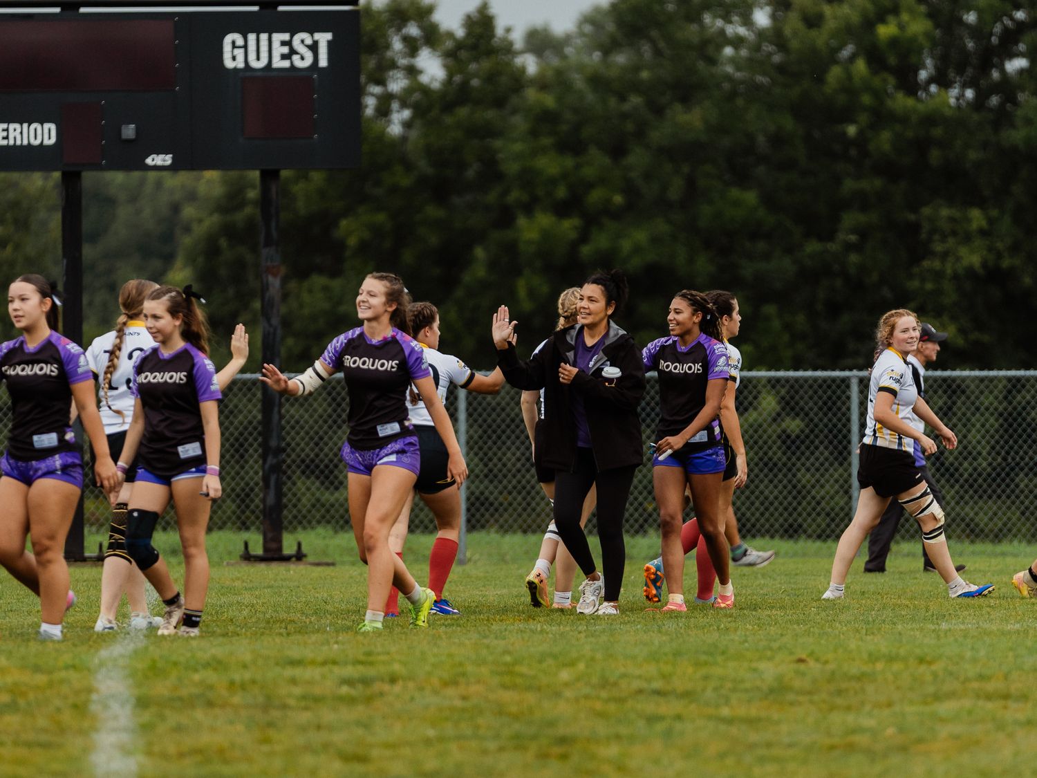 Women's rugby team celebrating on green field after game, wearing purple uniforms, with scoreboard visible in background.