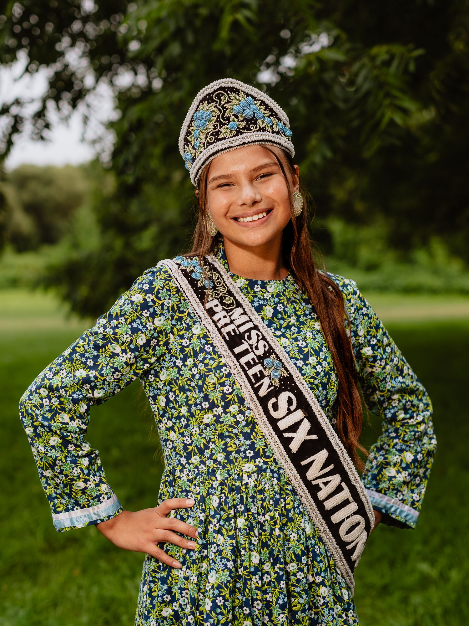 Young girl in floral dress wearing a decorative crown and Six Nations pageant sash, smiling outdoors.
