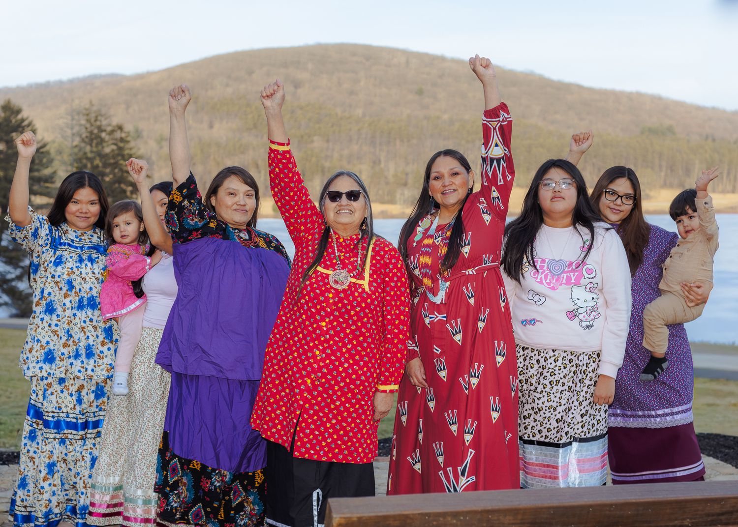 Group of Indigenous women raising fists in unity outdoors near a lake, wearing traditional and colorful clothing.