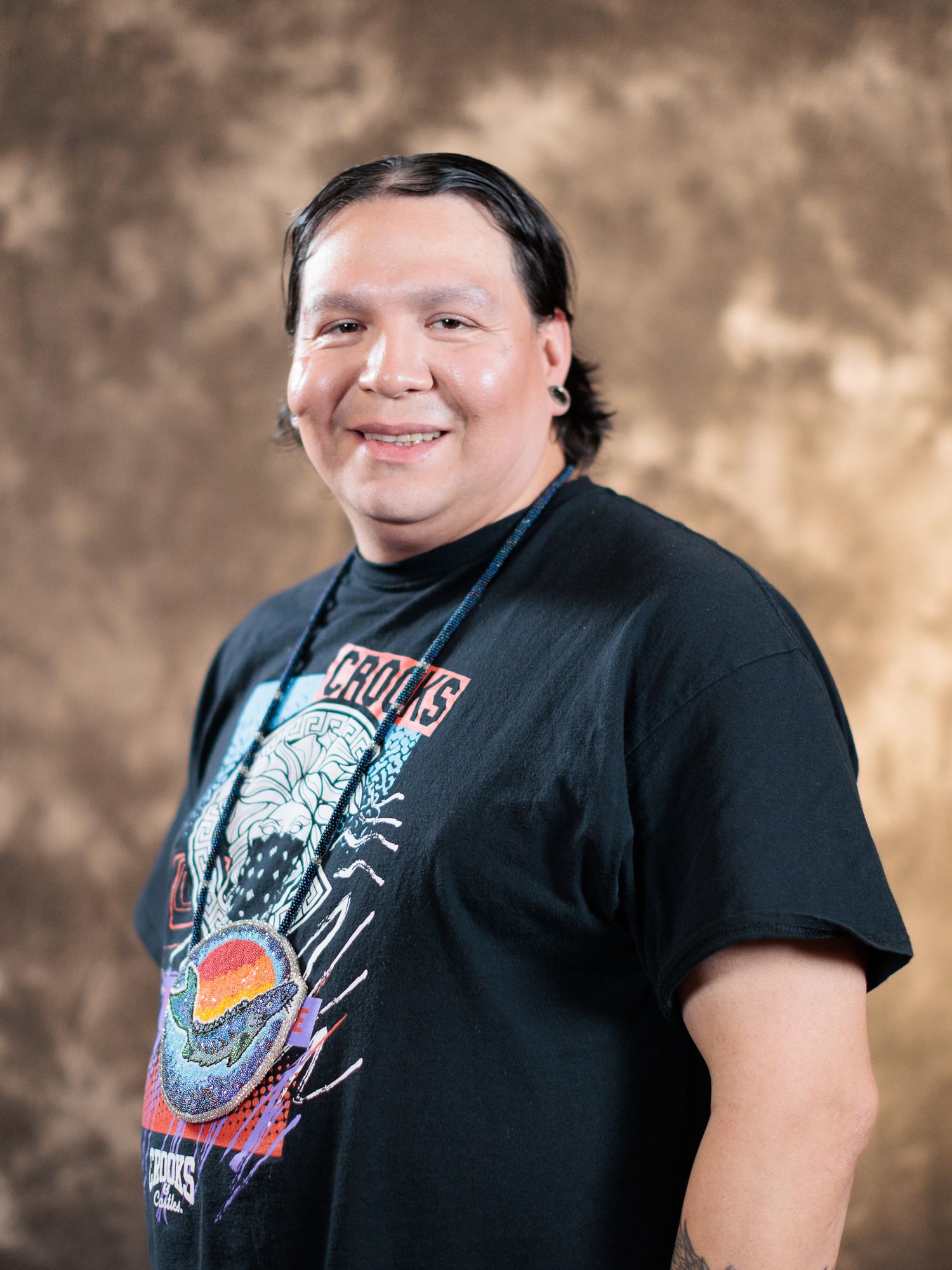 Man smiling in black graphic t-shirt posed against brown mottled studio backdrop.