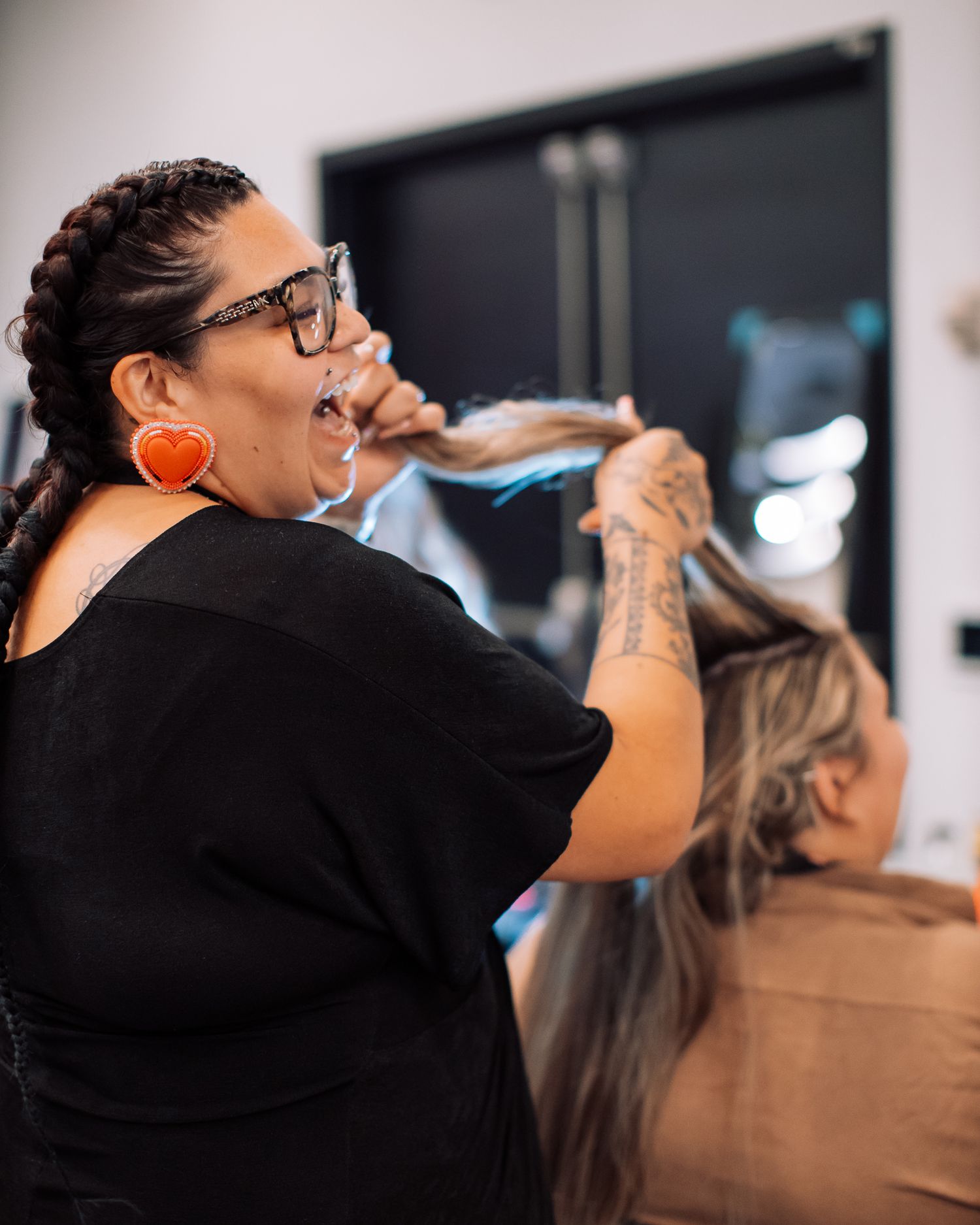Hairstylist with braids and orange earrings works on client hair in modern salon setting.