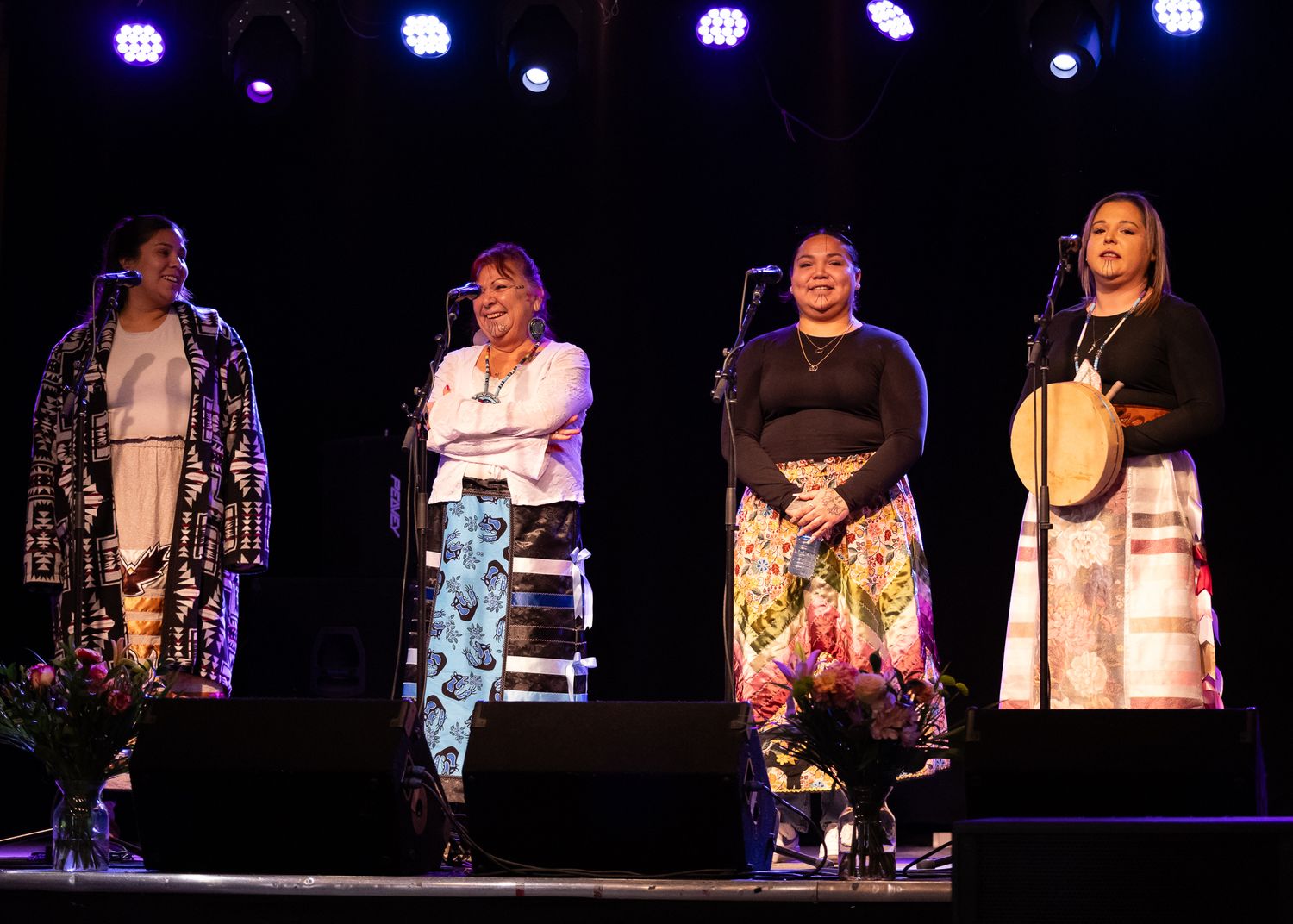Four Indigenous women performers on stage, one holding a drum, at a live music concert event.