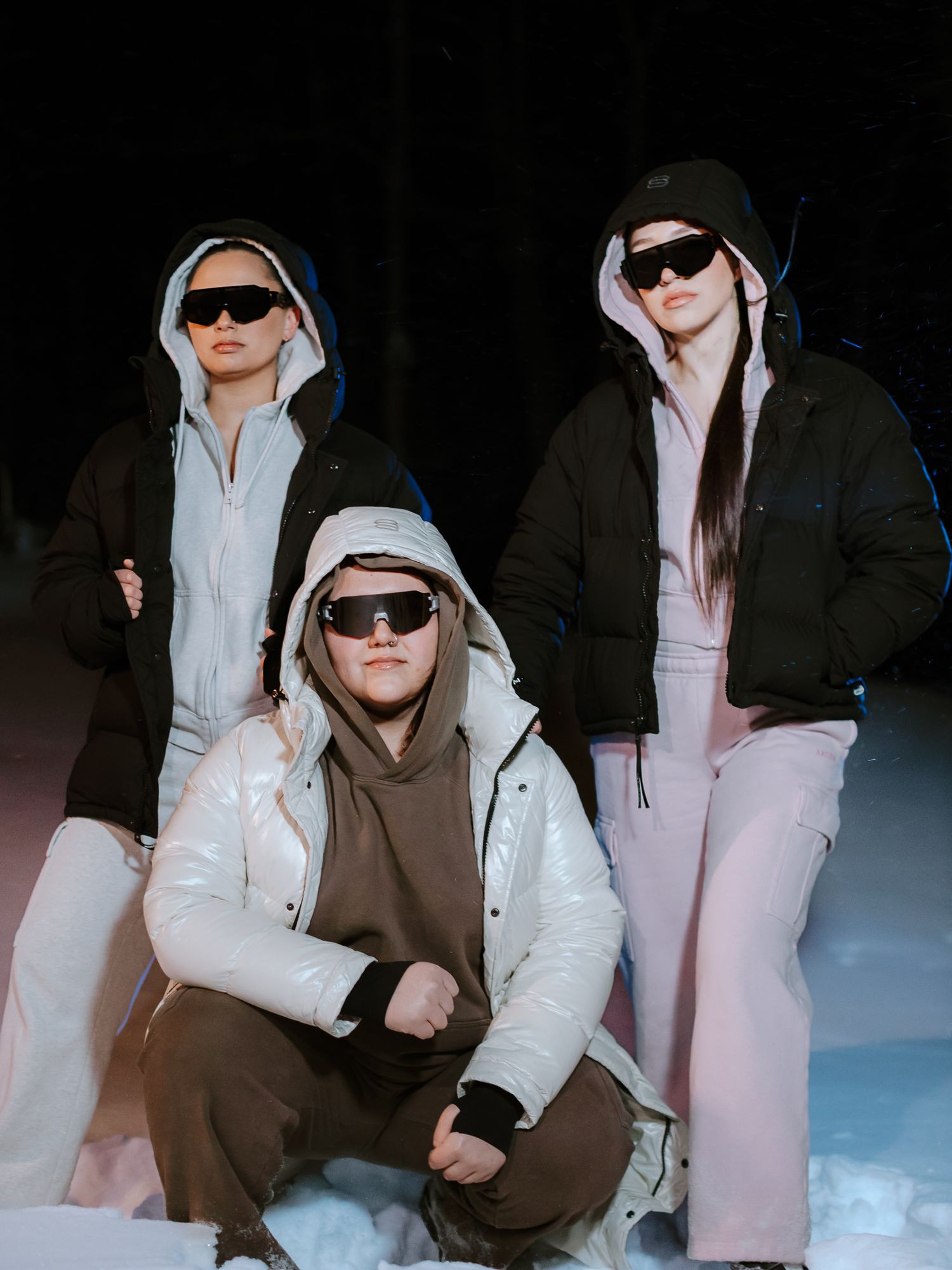Three women in hooded puffer jackets and sunglasses pose dramatically in snow at night.