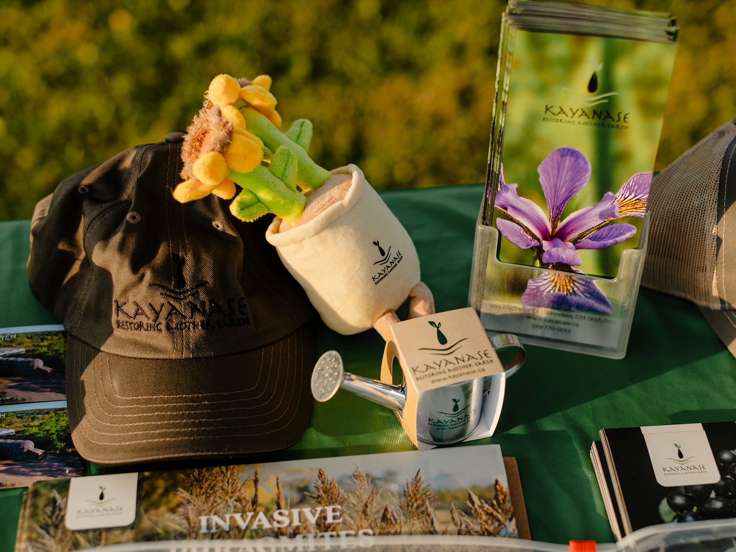 Kaynakii branded merchandise display featuring a hat, watering can, plant, and iris poster on a table with invasive species booklets.