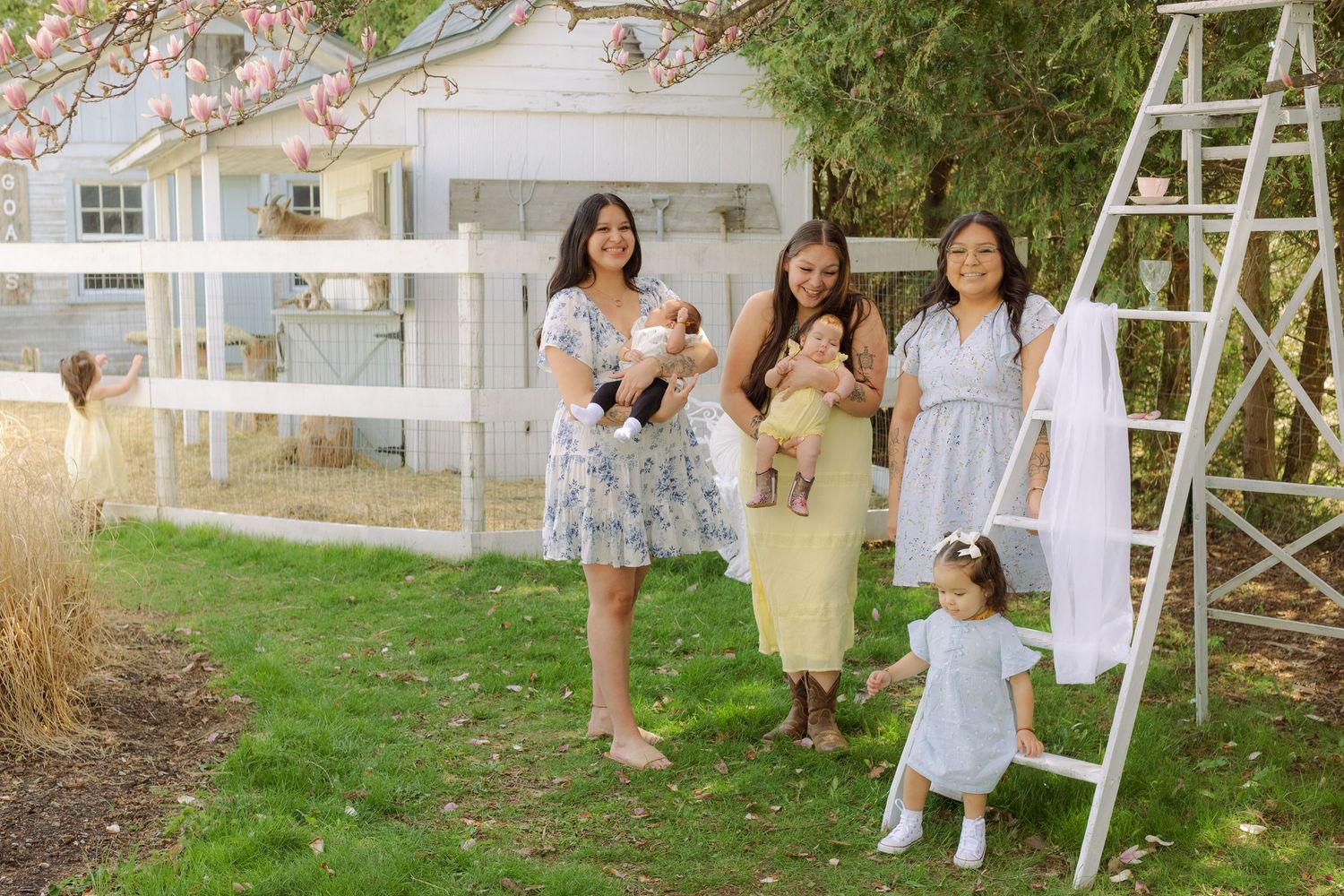 Four women in floral dresses pose outdoors near a white fence and ladder, holding babies on a sunny spring day.