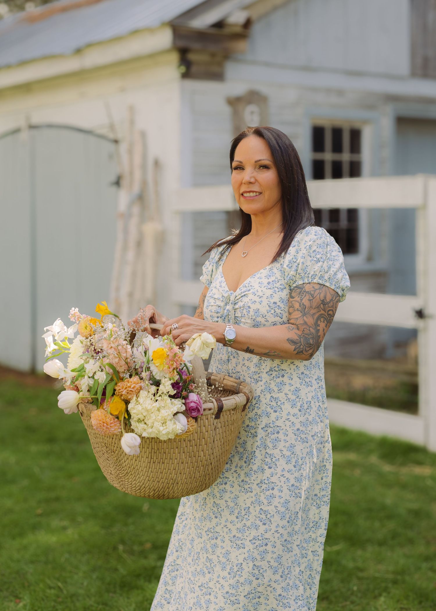 Woman in floral dress holding a basket of colorful flowers outside a rustic barn on a sunny day.
