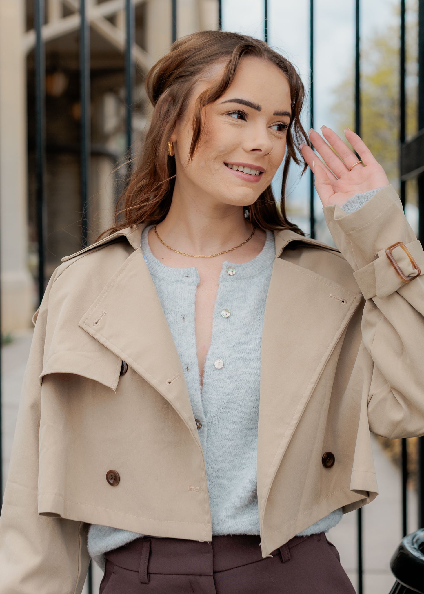 Young woman in beige trench coat smiling and waving outdoors near iron gate.