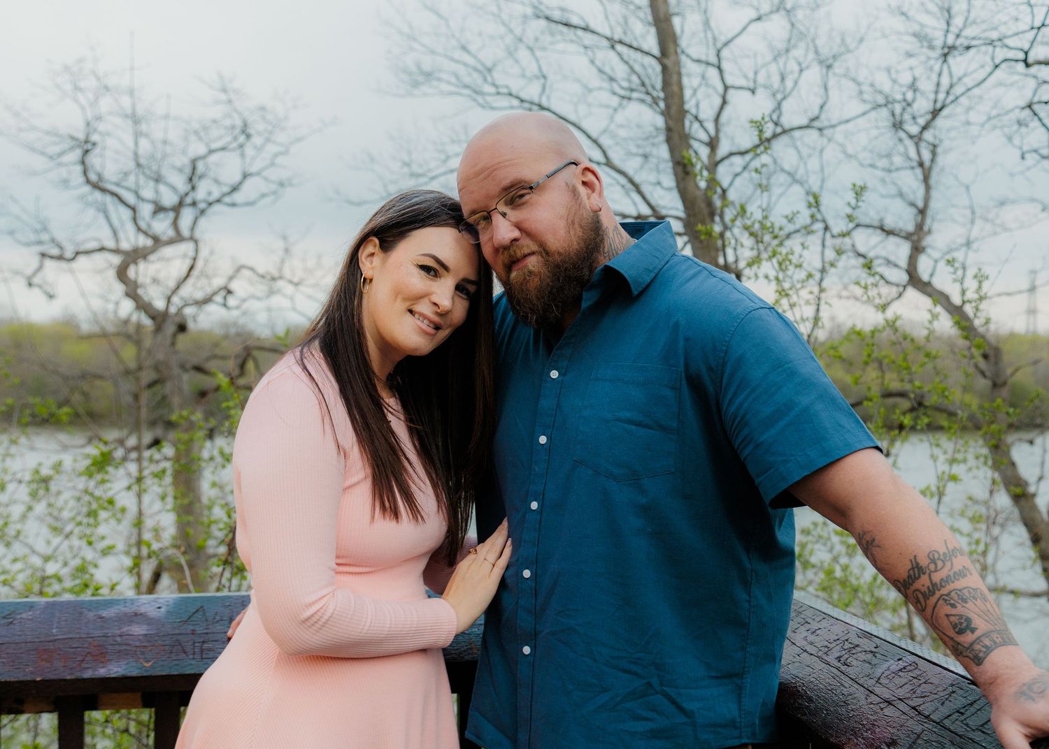 Couple posing outdoors near a lake, woman in pink dress leaning on man in blue shirt, trees in background.